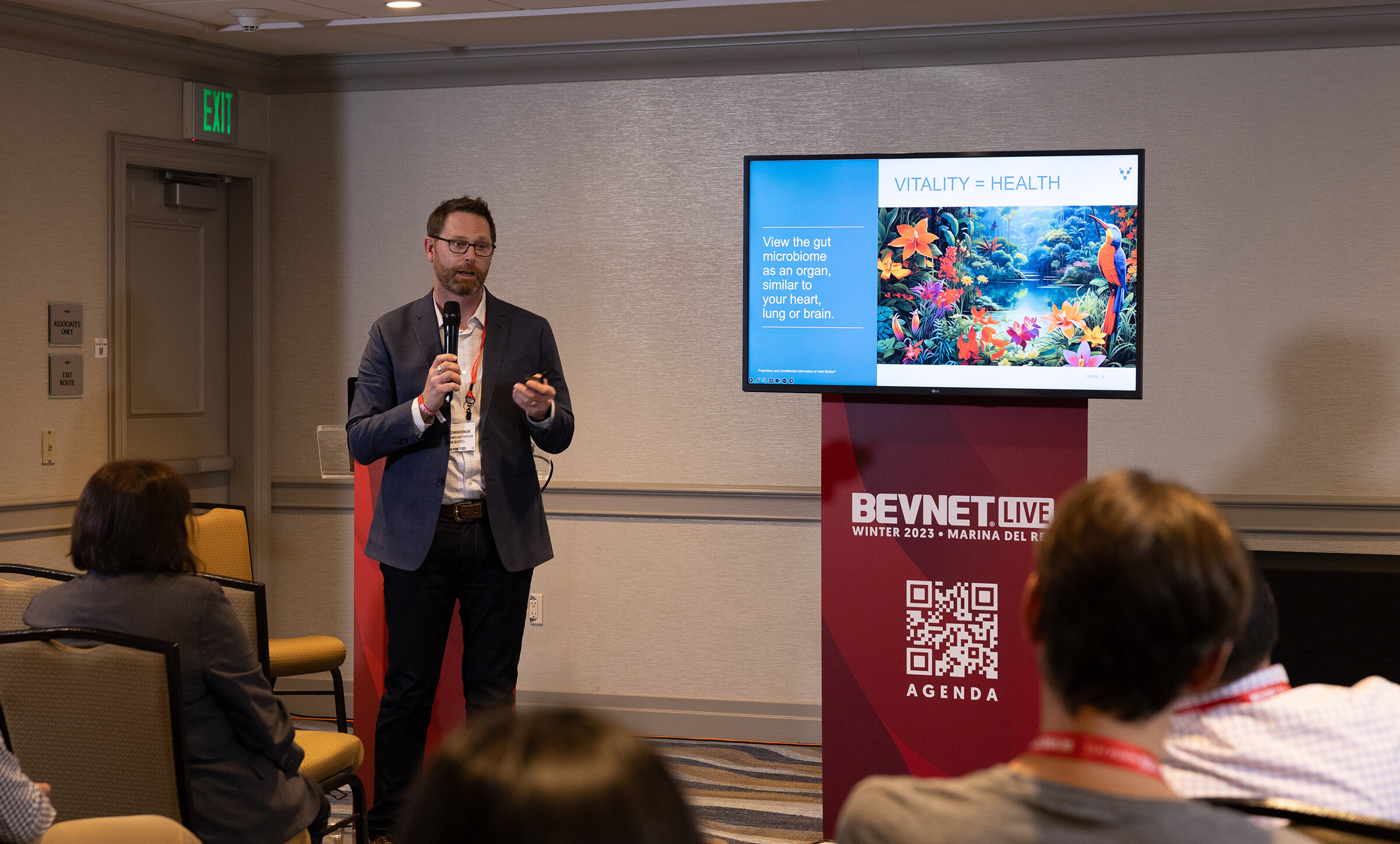 A man in a dark suit stands at a lectern marked BevNET Live next to a screen, with attendees in the foreground.