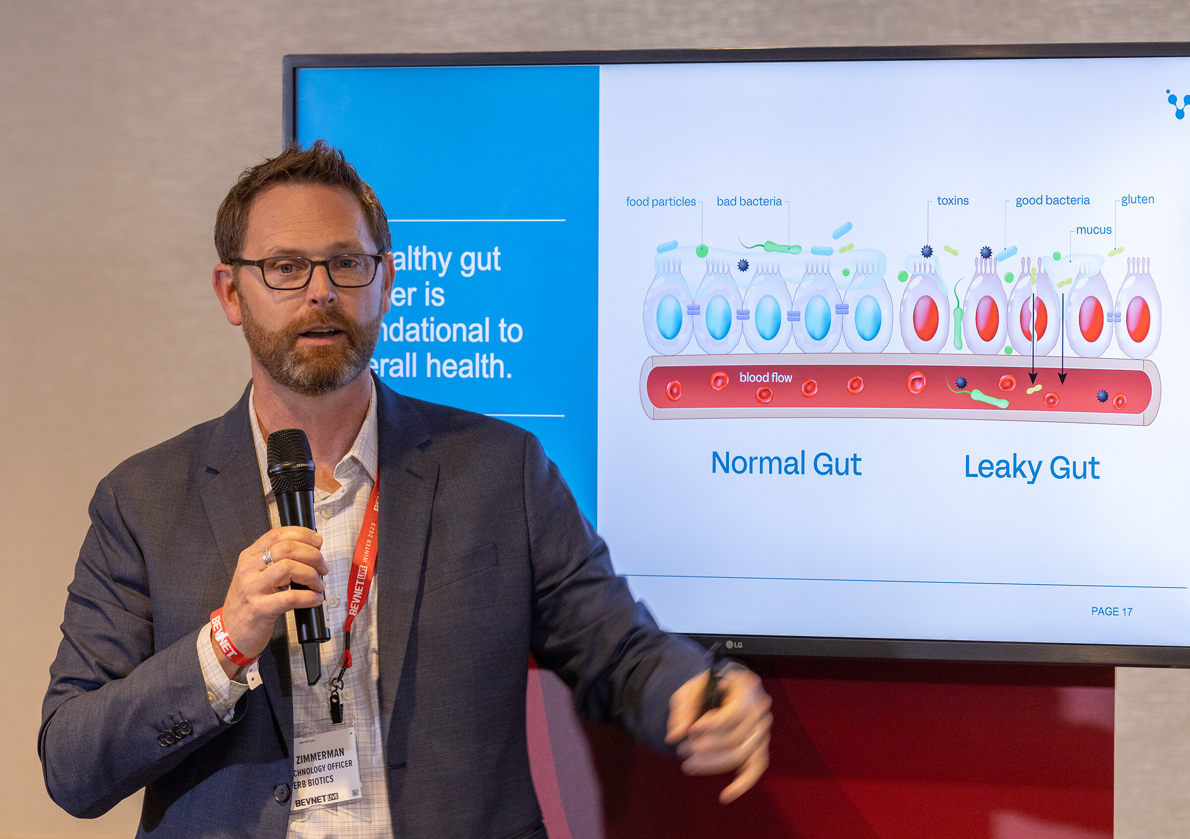 A man with a beard and glasses holds a microphone in front of a slide about normal and leaky gut.