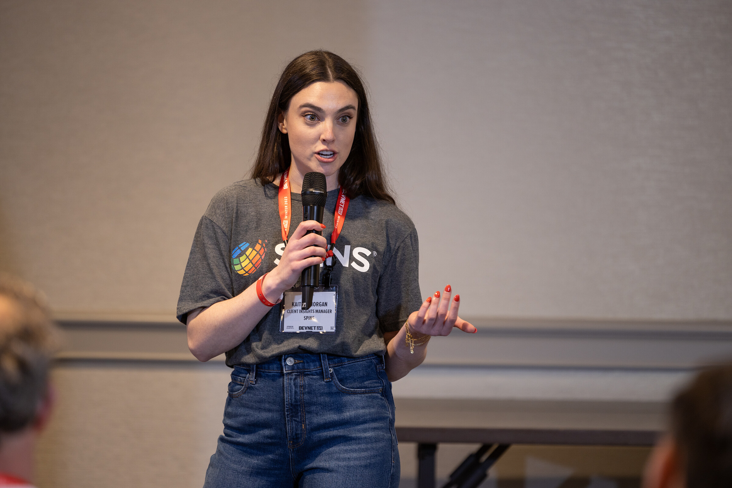 A woman in a SPINS T-shirt and jeans speaks into a microphone in a meeting room.