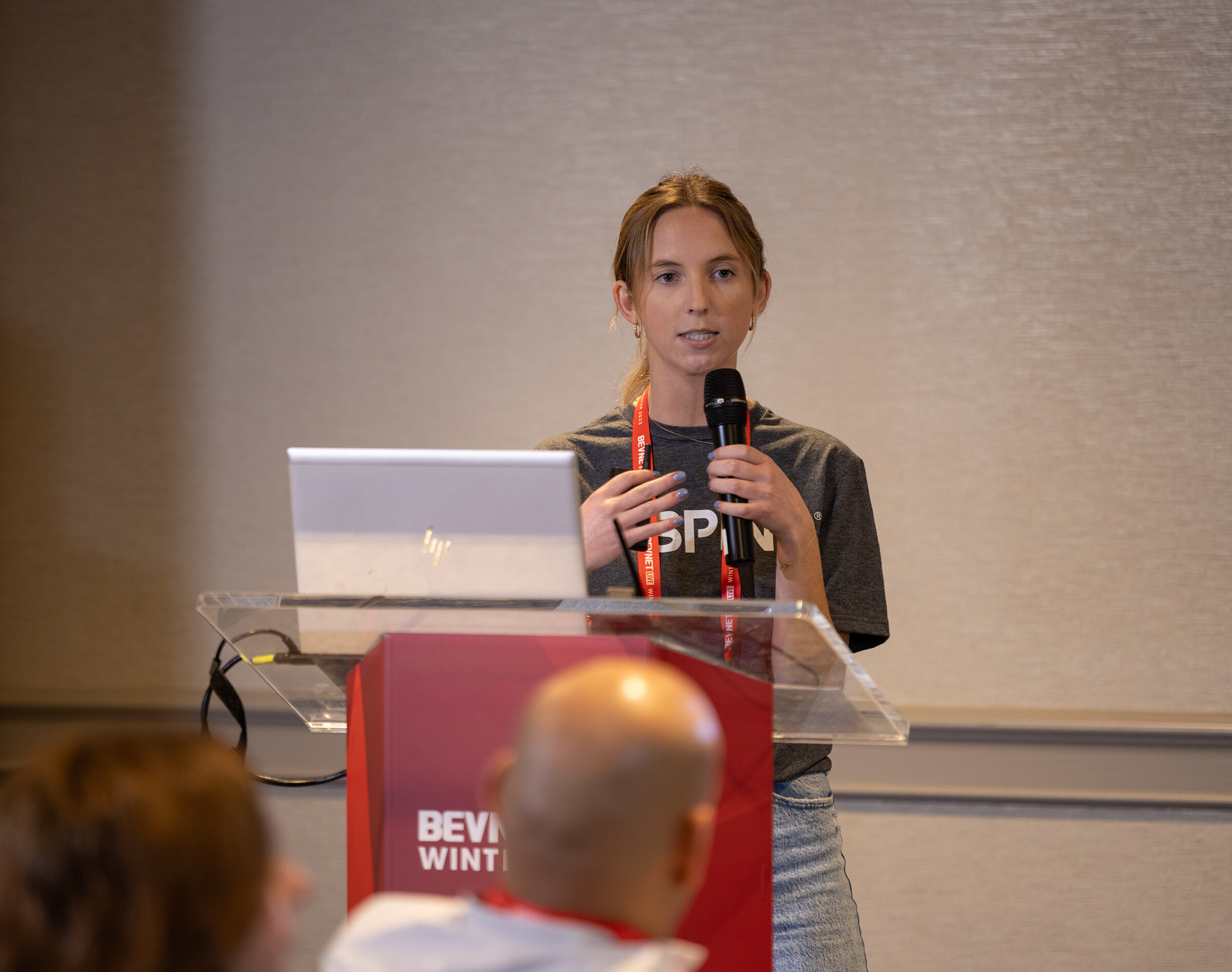 A woman in a gray SPINS T-shirt speaks from a lectern with a laptop while attendees sit in the foreground.