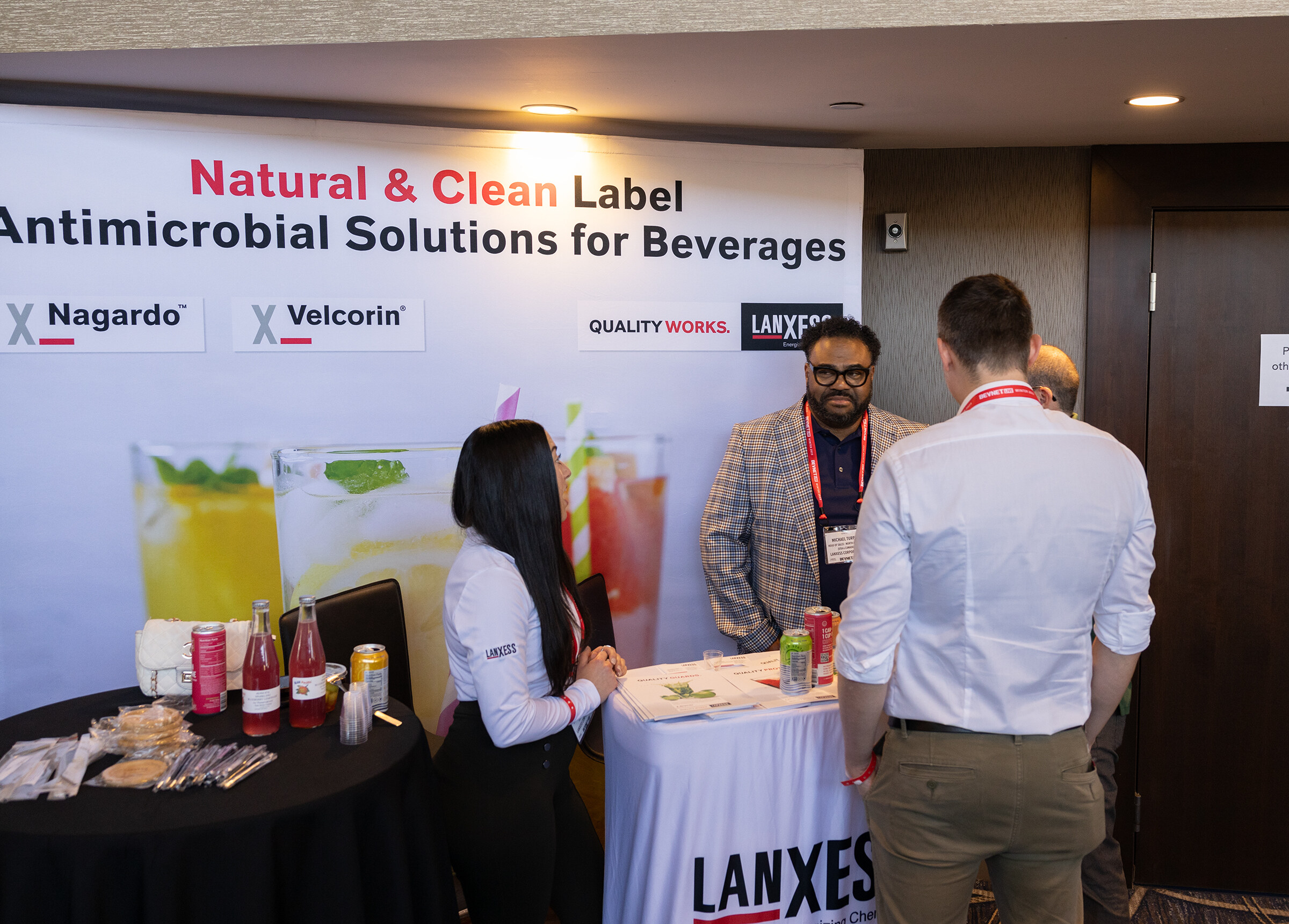 A table with a Lanxess tablecloth and pitchers of drinks, with exhibitors talking to two attendees.