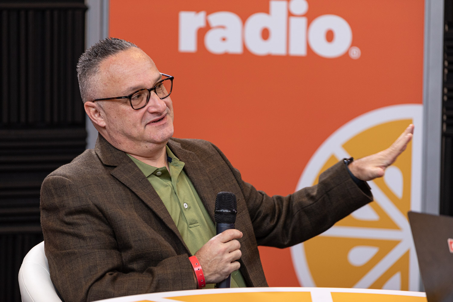 A man in a brown blazer and green shirt speaks into a microphone in front of a Taste Radio backdrop.