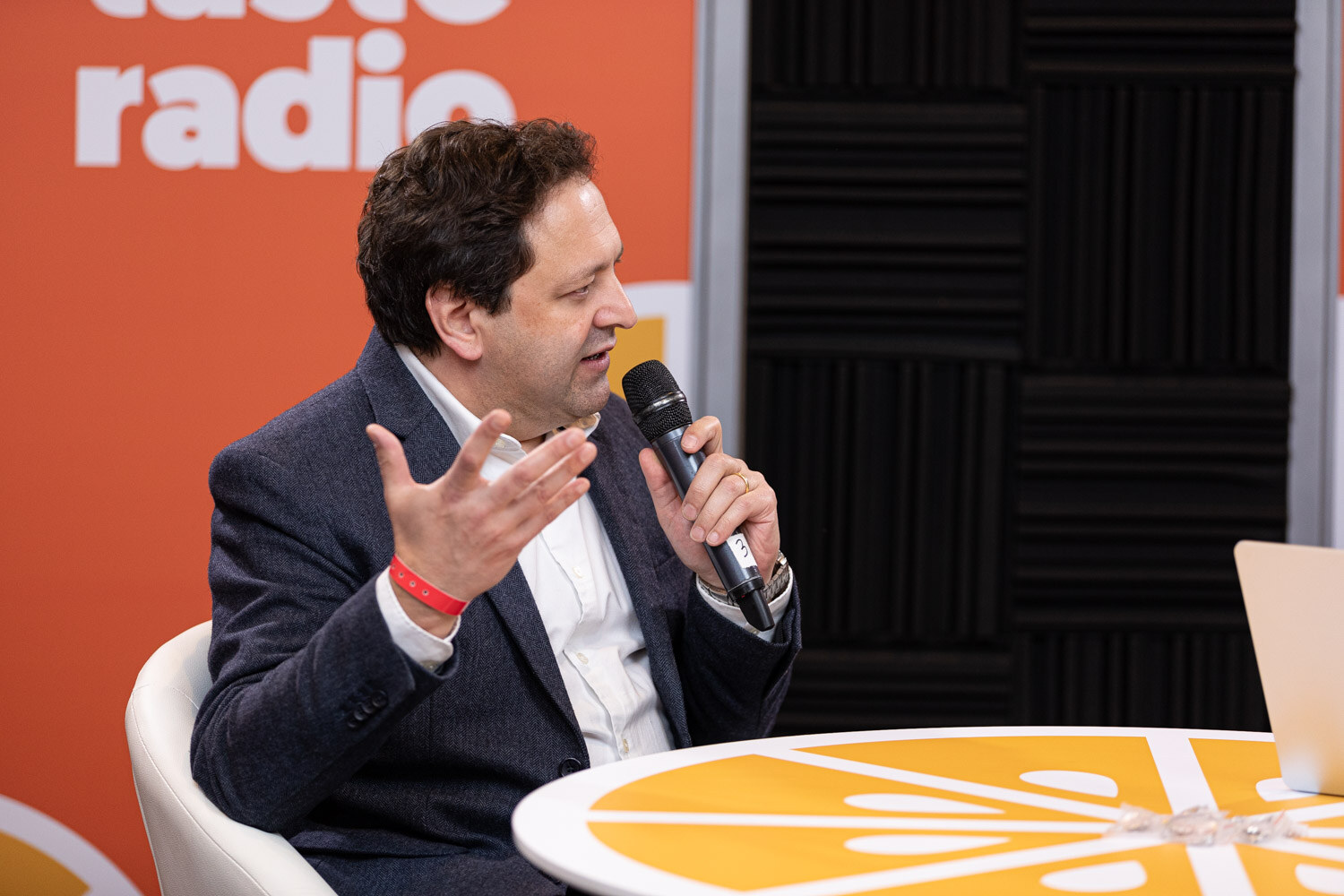 A man with dark curly hair in a dark suit holds a microphone and gestures.