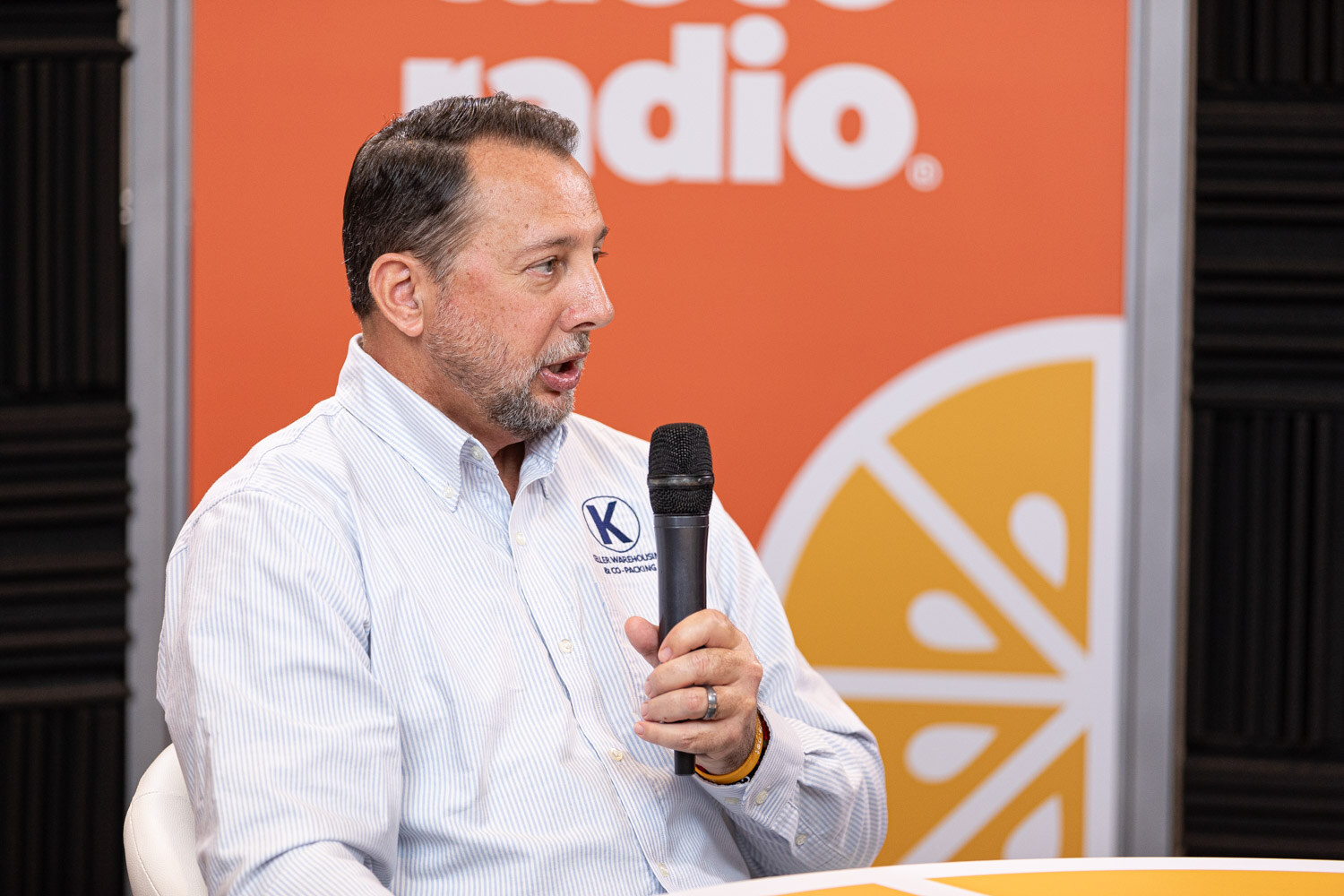 A man in a white shirt with a K logo holds a microphone in front of the Taste Radio backdrop.