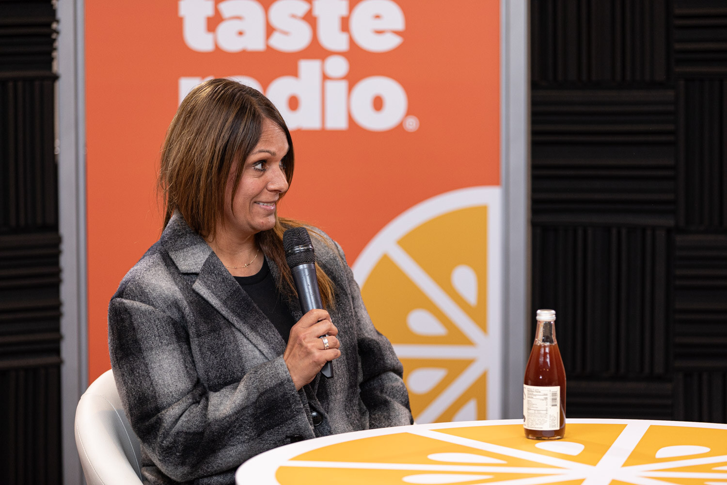 A woman in a gray plaid jacket holds a microphone with a bottle on the table.