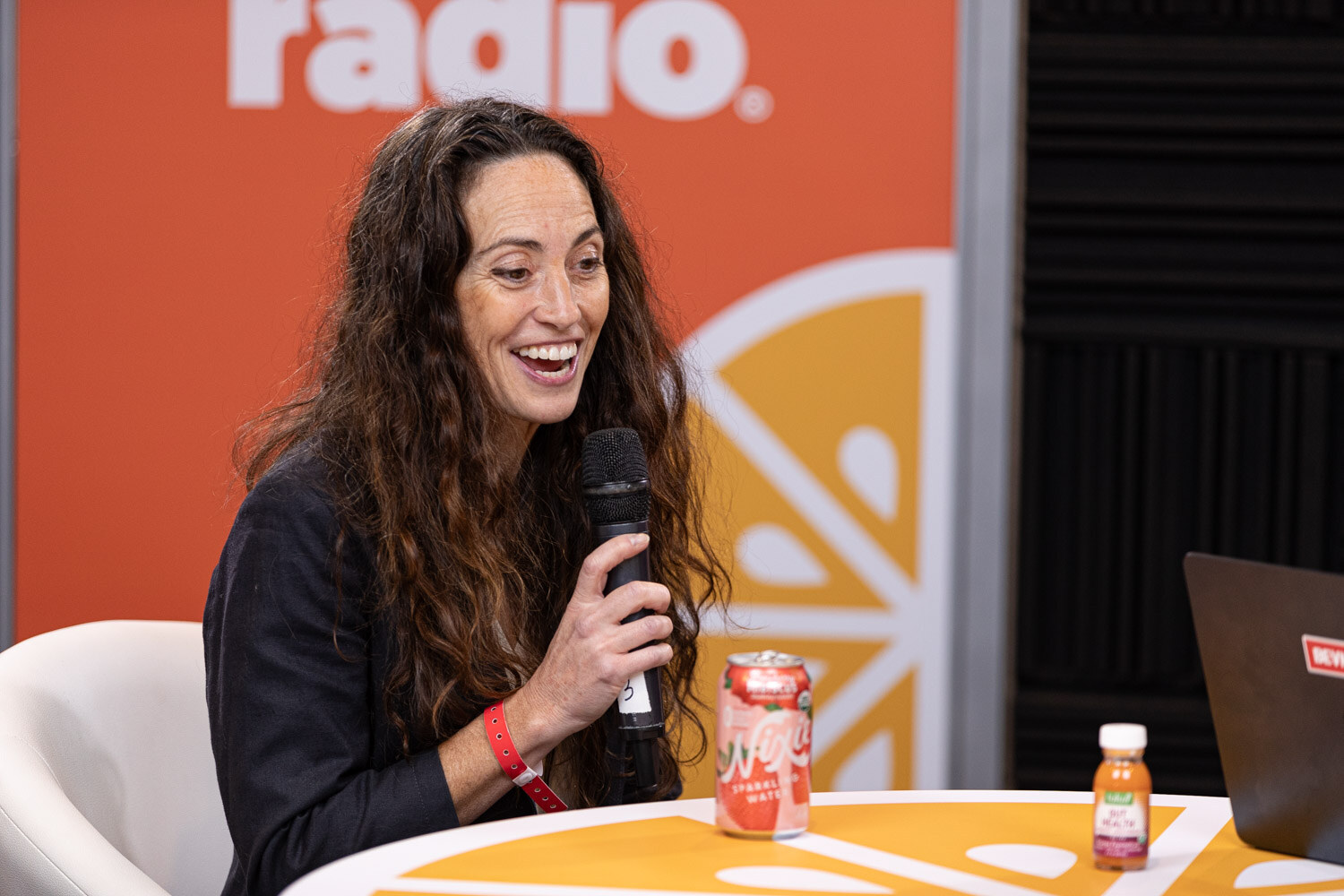 A woman with long dark hair and a lanyard laughs while holding a microphone, with cans on the table.