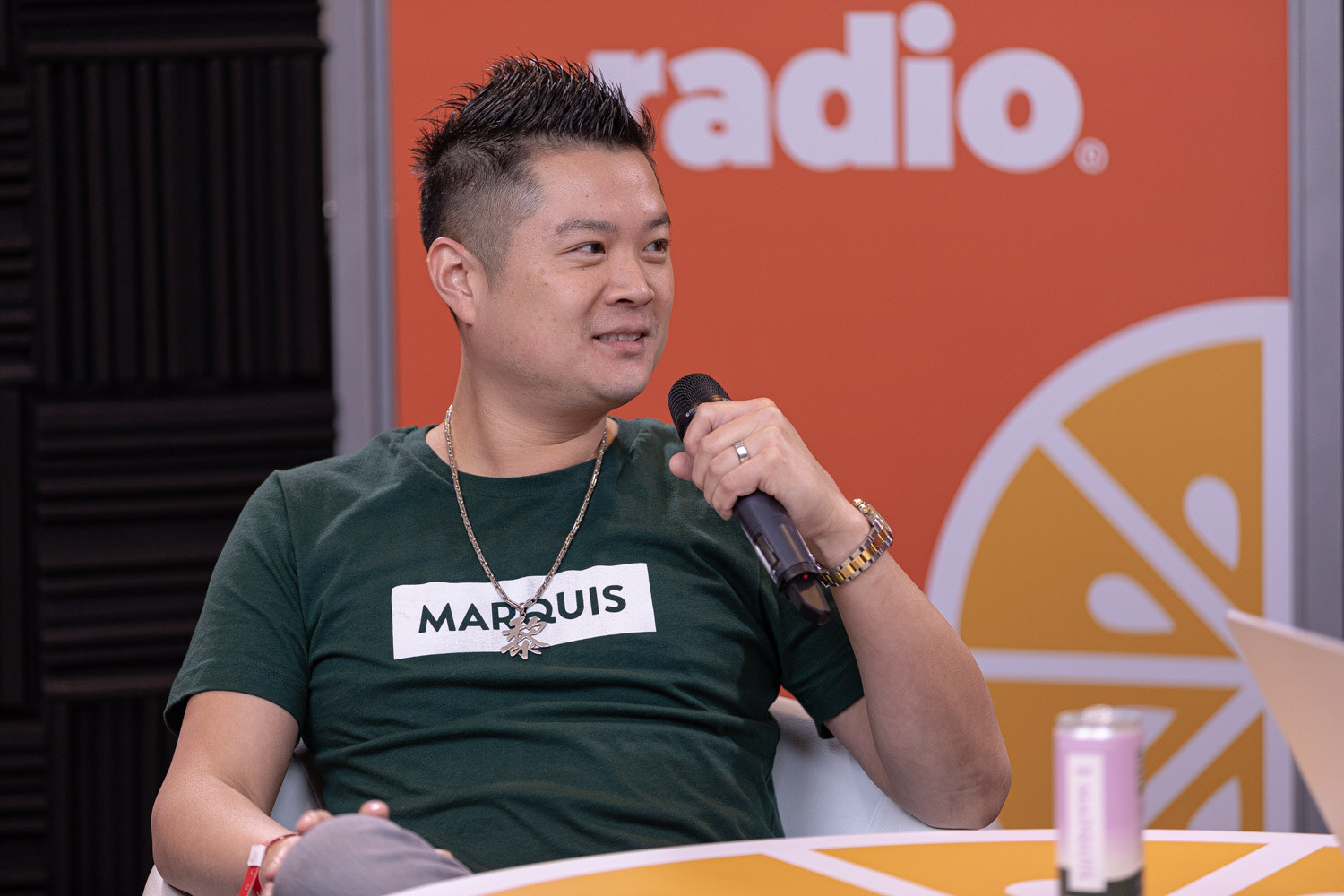 A man in a green Marquis T-shirt looks to the side while holding a microphone.