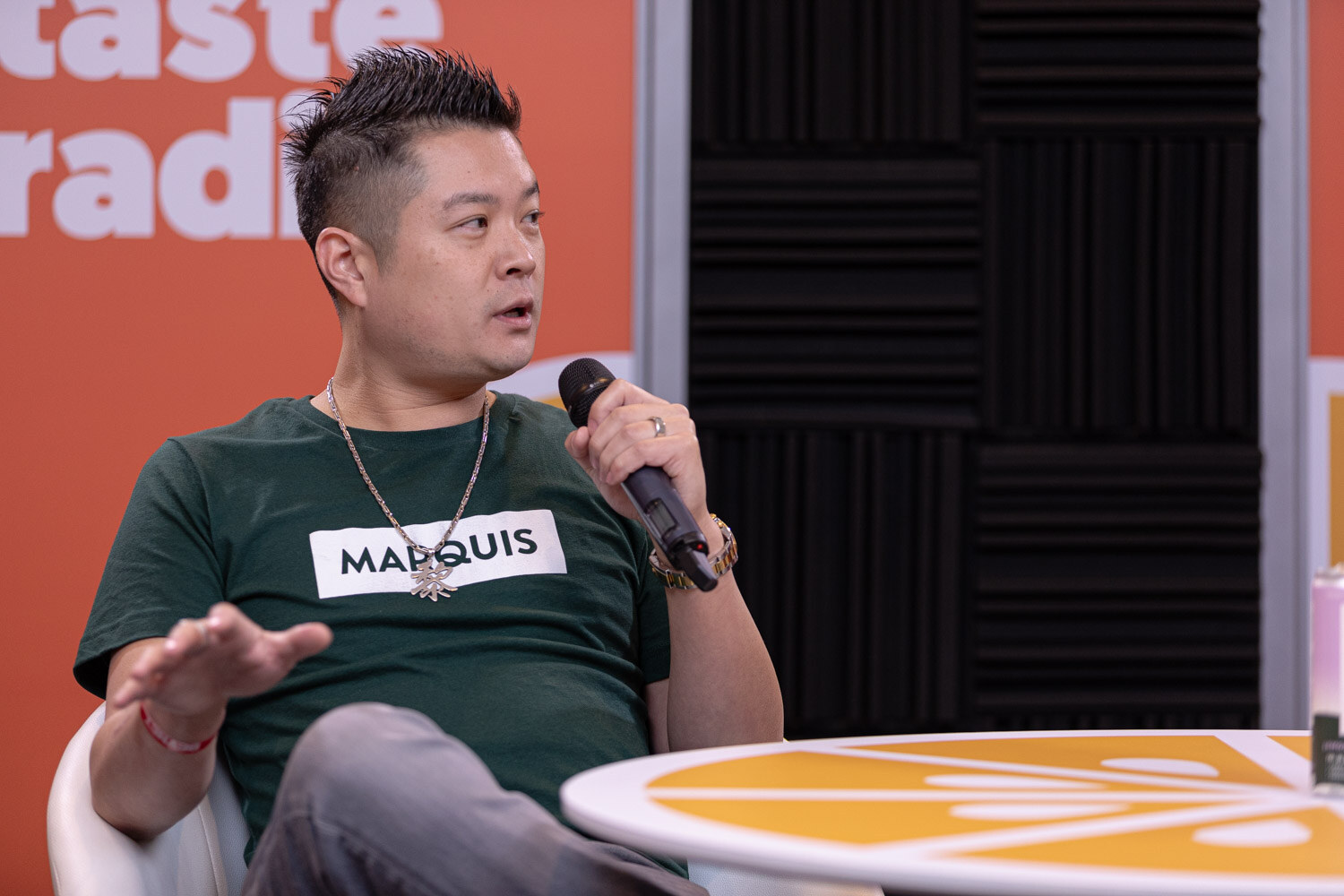 A man in a green Marquis T-shirt gestures with his left hand while holding a microphone.