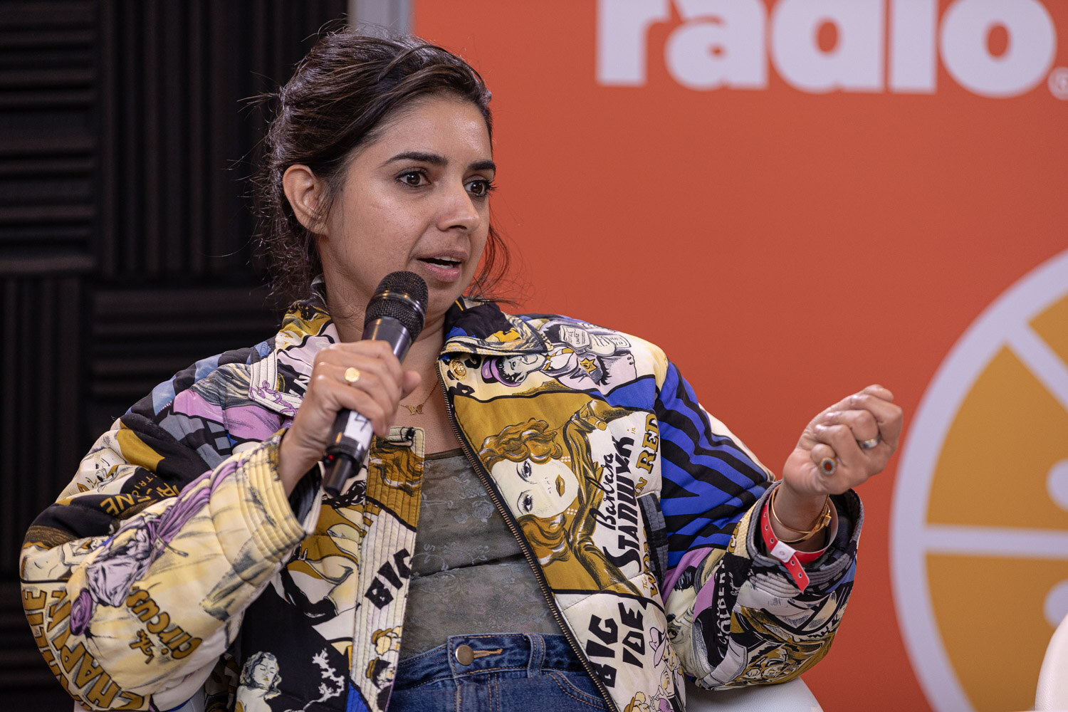 A woman in a colorful patterned jacket holds a microphone and gestures.