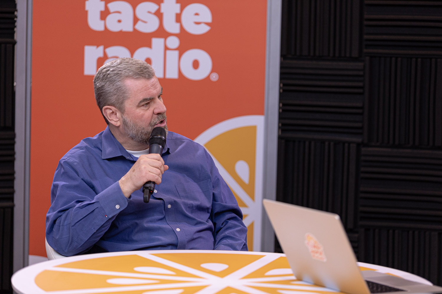 A man in a blue button-up shirt speaks into a microphone in front of an orange Taste Radio backdrop.
