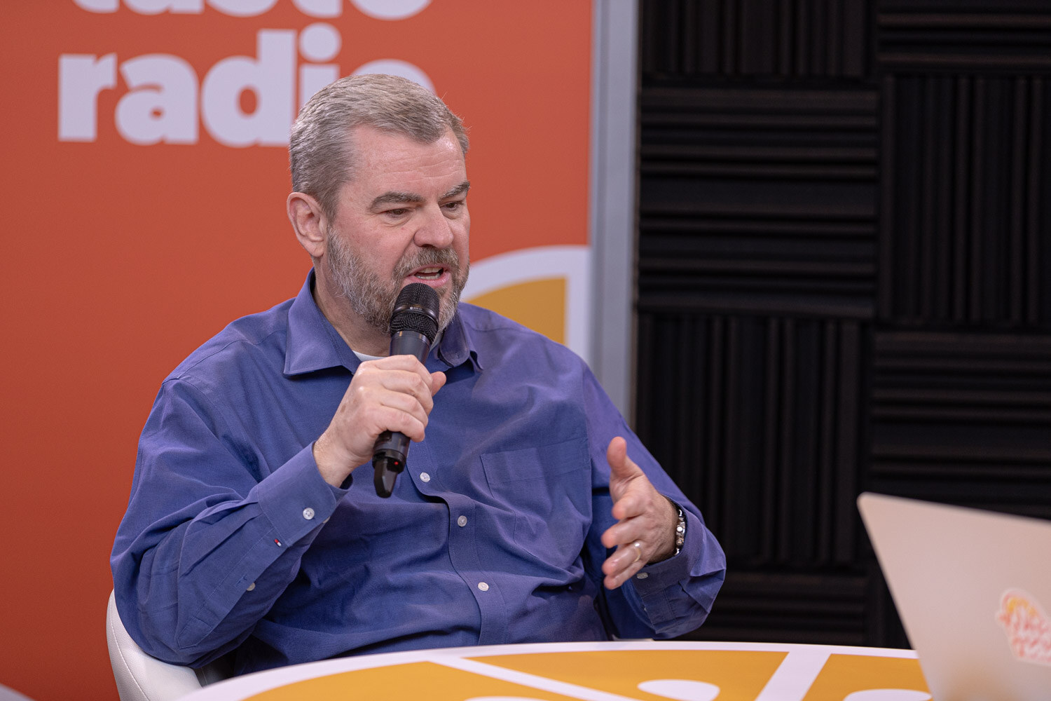 A man in a blue shirt speaks into a microphone in front of a Taste Radio backdrop.