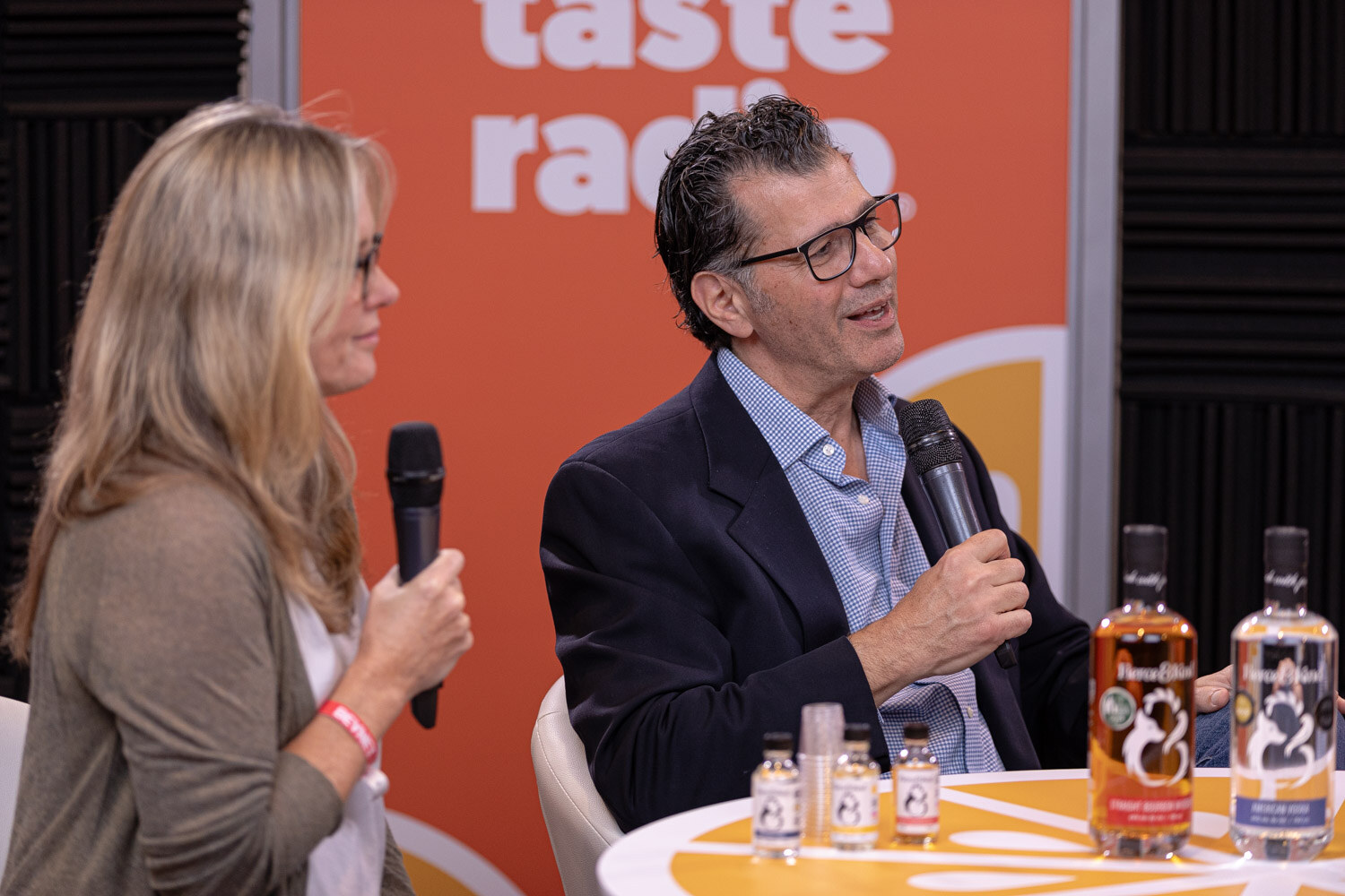 A woman and a man in a dark suit speak into microphones with two bottles on a table in a Taste Radio studio.