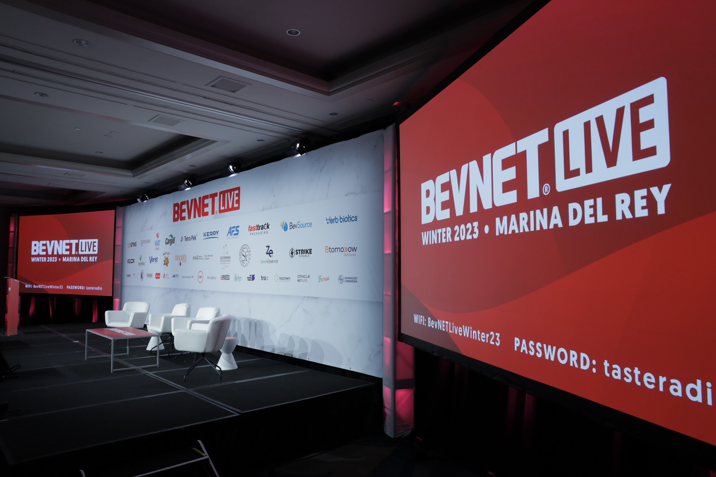 A stage with white lounge chairs, a large red screen reading BevNET Live Winter 2023 Marina del Rey and a sponsor backdrop.