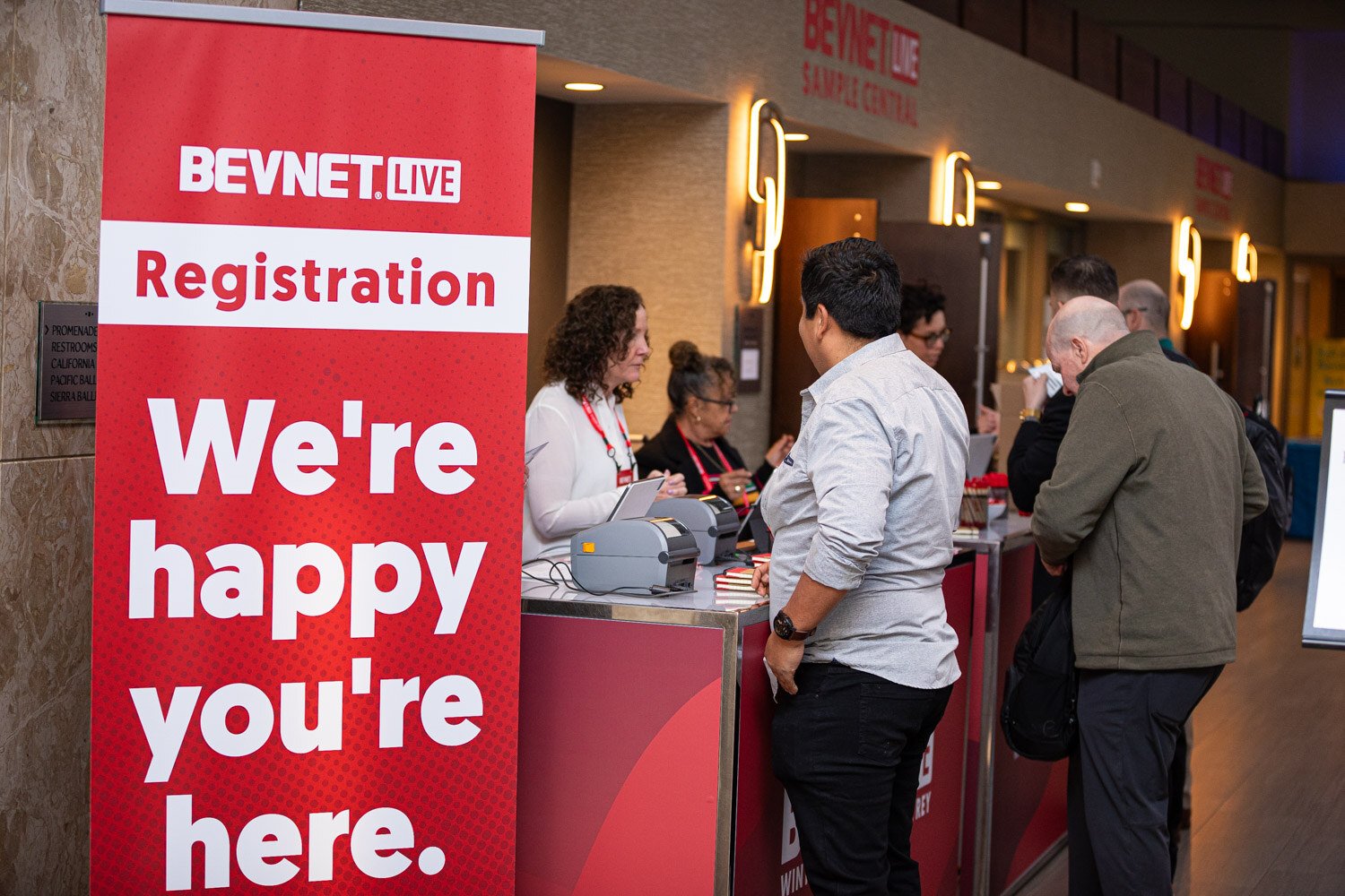 Attendees stand at a registration counter beside a tall red banner that says We're happy you're here.