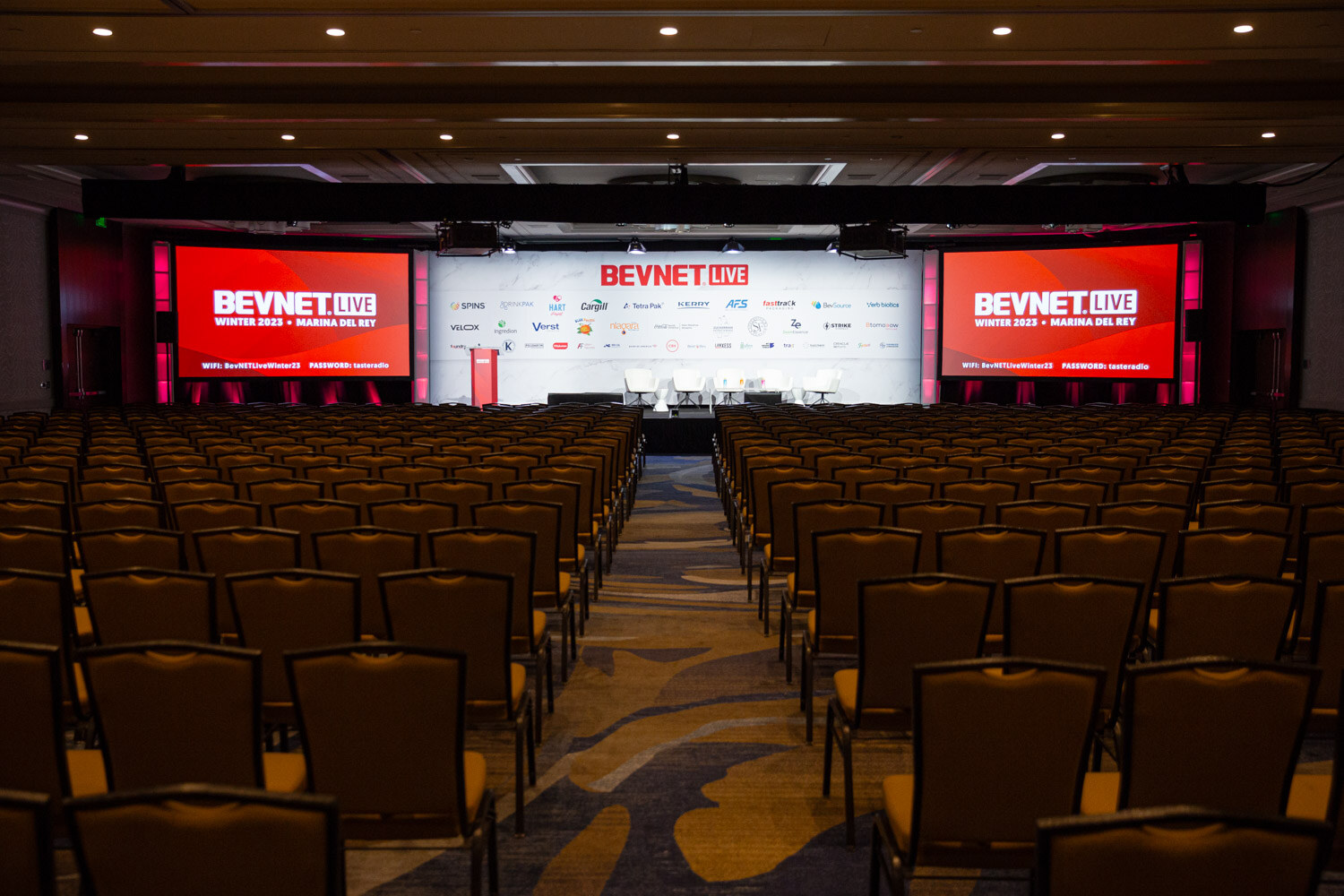 A wide view of rows of chairs facing a stage with large red BevNET Live screens.