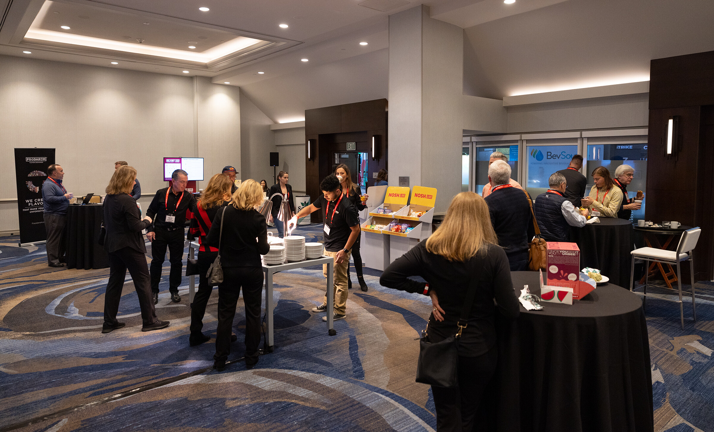 People stand around black-covered tables in a hotel meeting space with window light at the back.