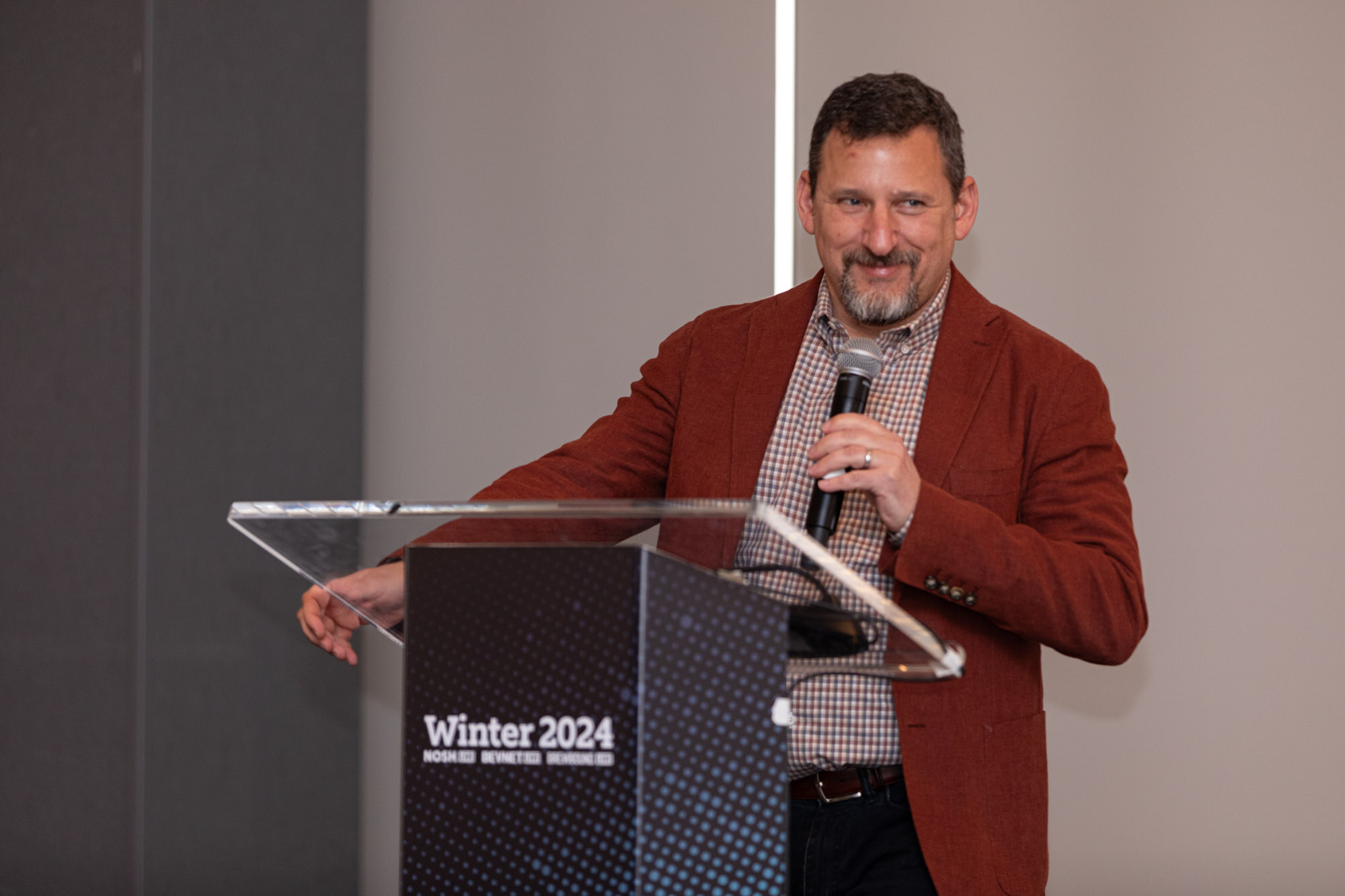 Smiling man in a red blazer and checked shirt holds a microphone at a clear lectern with a Winter 2024 sign.