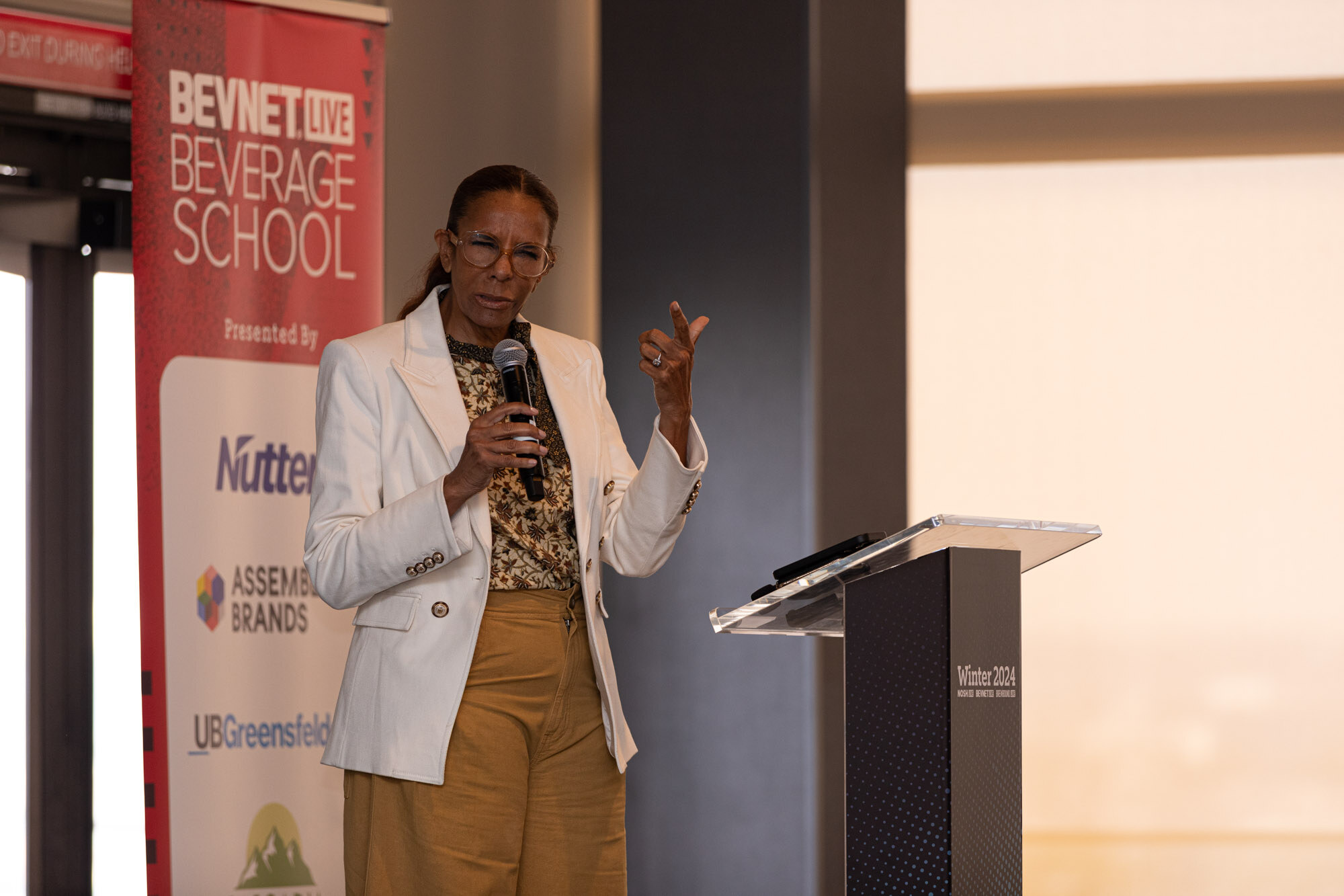 Woman in a white blazer holds a microphone and gestures beside a red BevNET Live Beverage School banner.