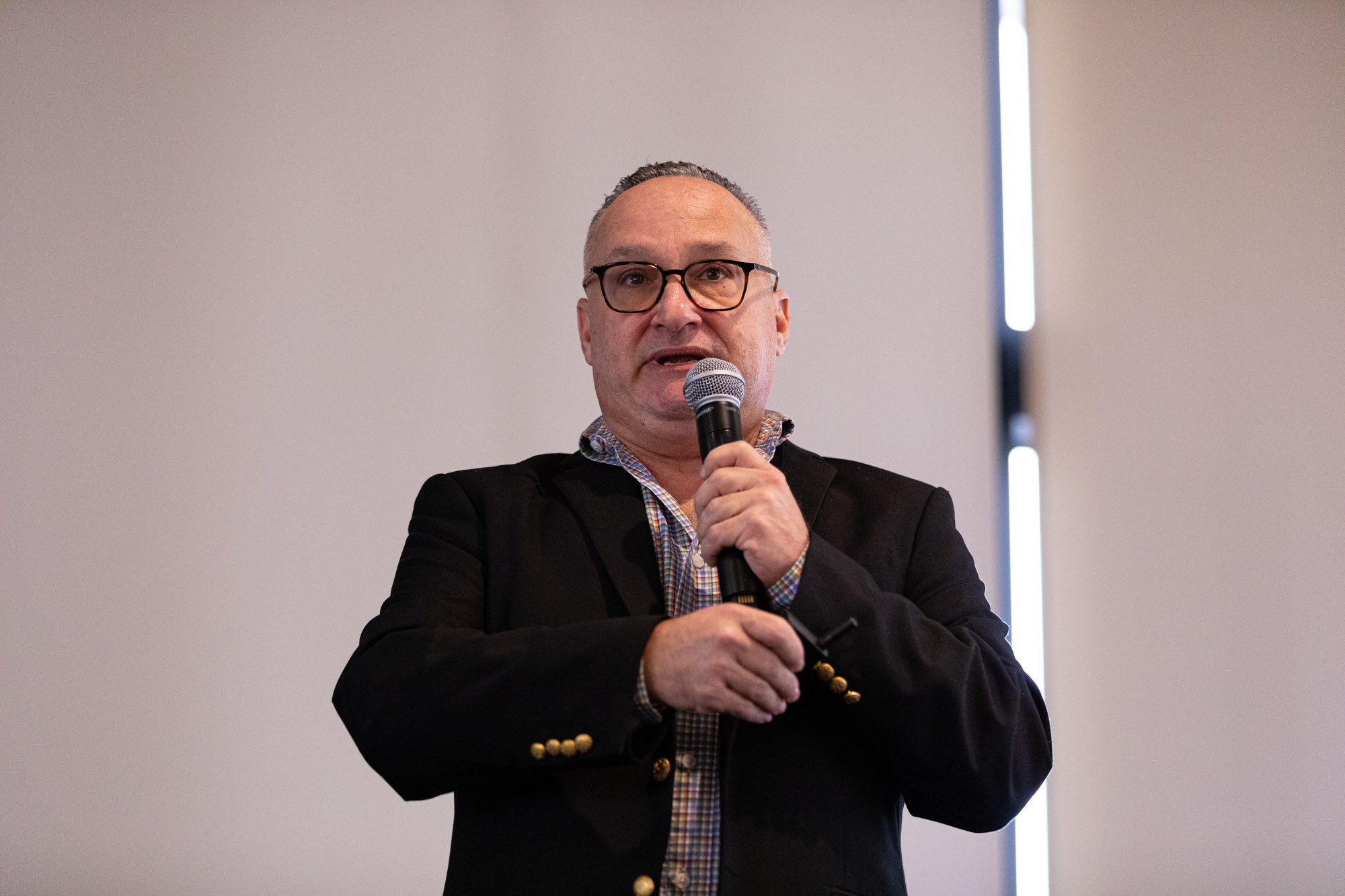 Man with glasses in a dark blazer holds a microphone in front of a plain gray wall.