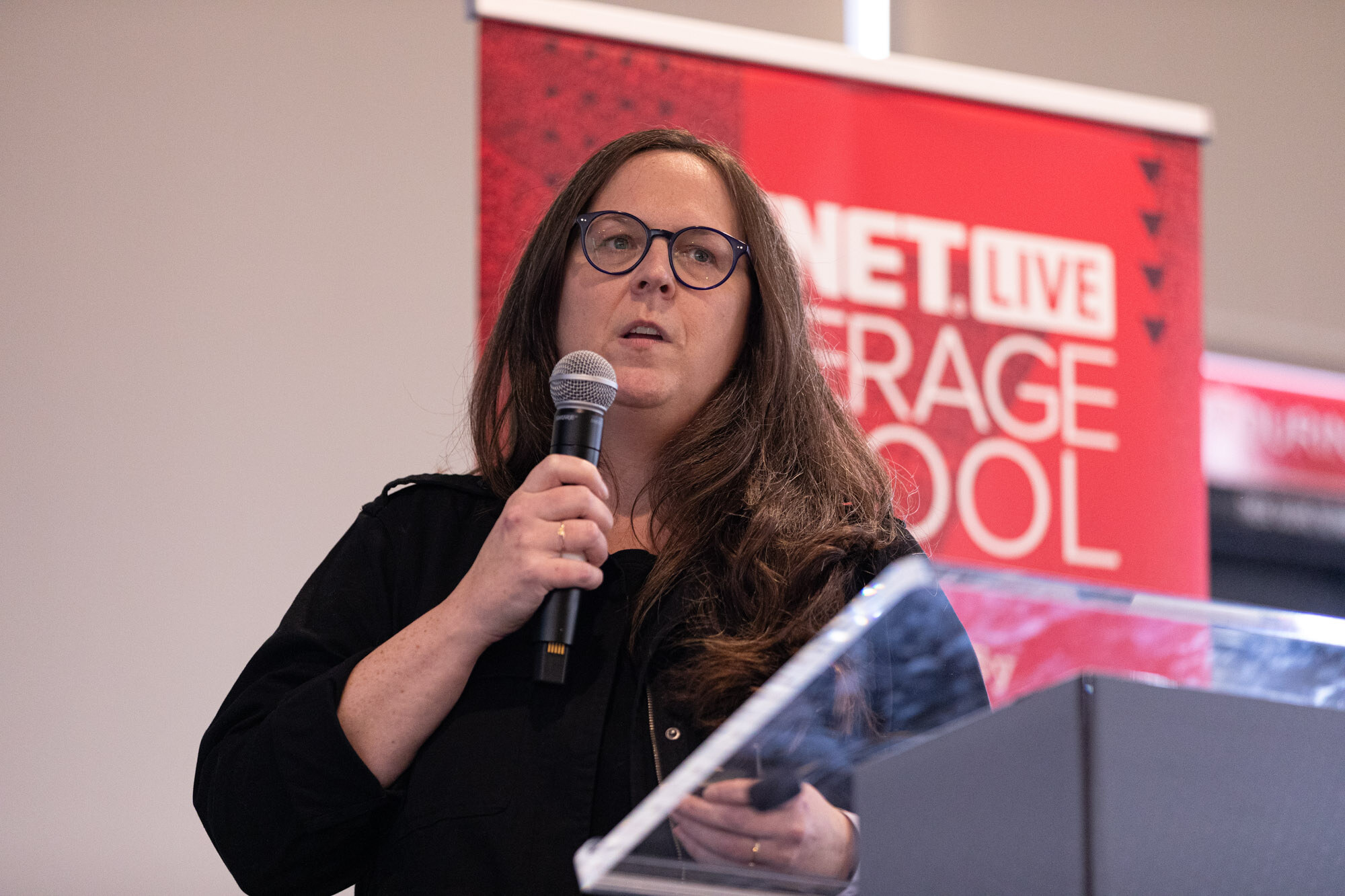 Woman with long brown hair and glasses speaks into a microphone at a clear lectern with a BevNET Live Beverage School banner.
