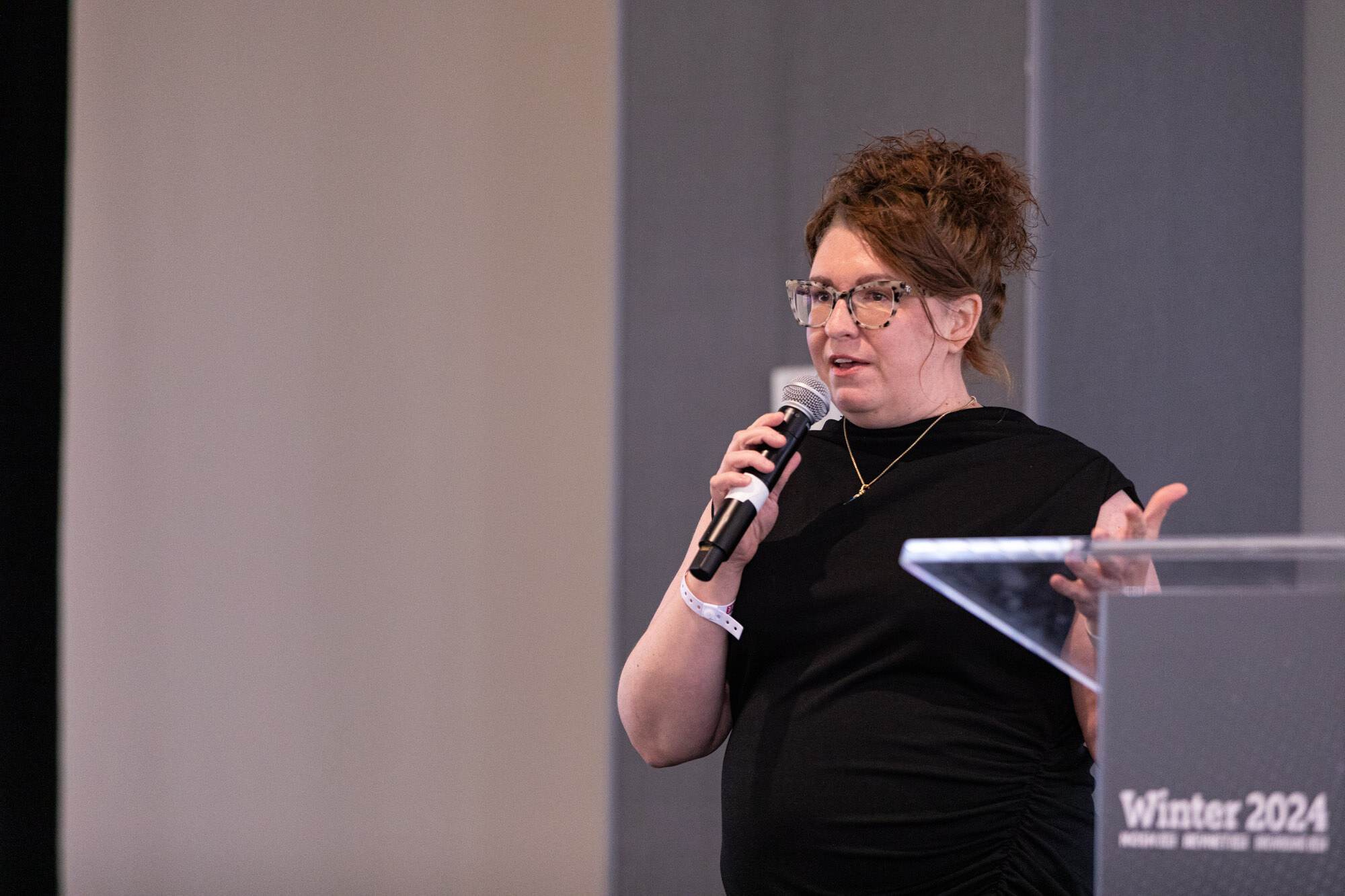 Woman in a black dress speaks into a handheld microphone beside a Winter 2024 lectern.