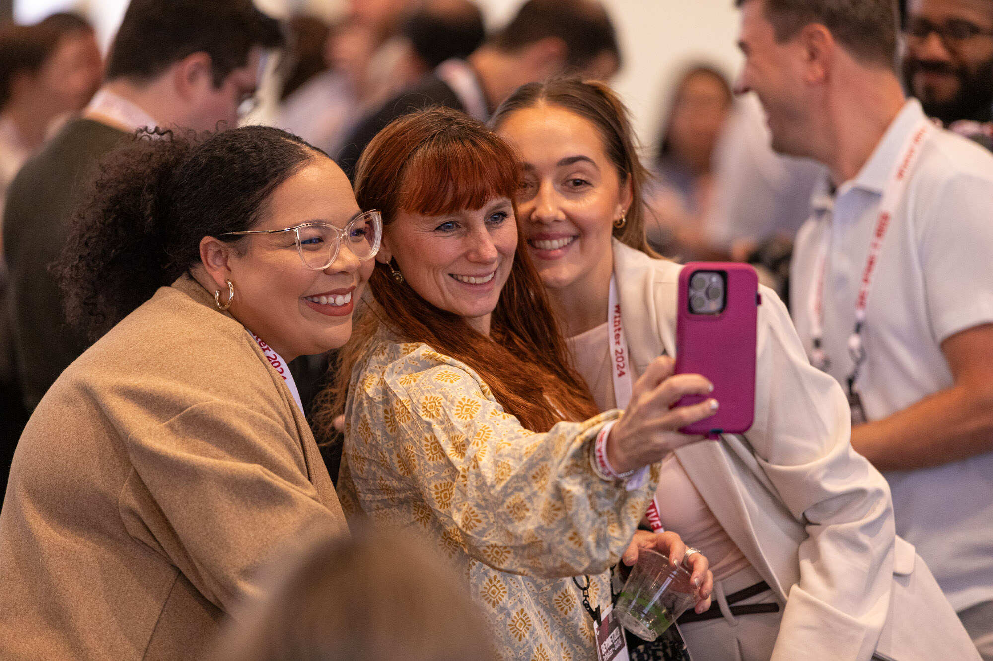 Three smiling women crowd together for a selfie with a magenta phone while other attendees stand behind them.