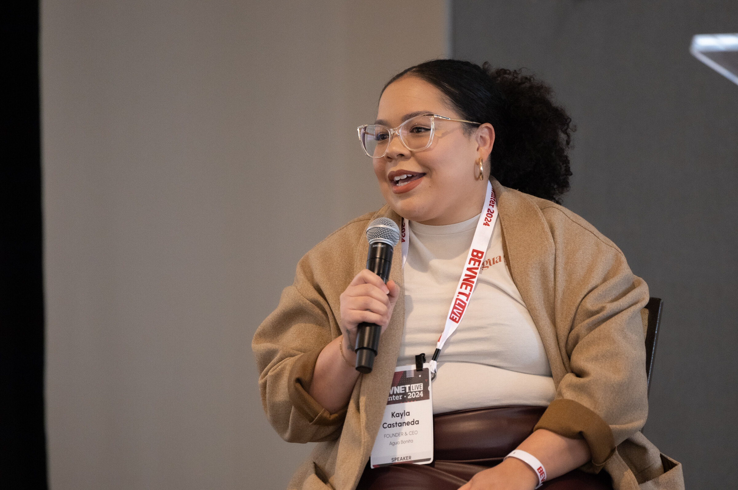 Woman in a camel coat holds a microphone and speaks while seated, wearing a BevNET Live lanyard.