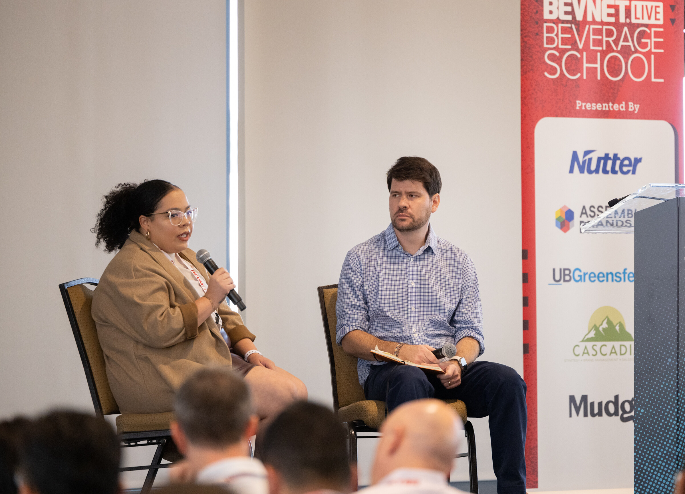 Two people sit in chairs on a small stage beside a red Beverage School banner listing sponsors including Nutter and UB Greenfelder.