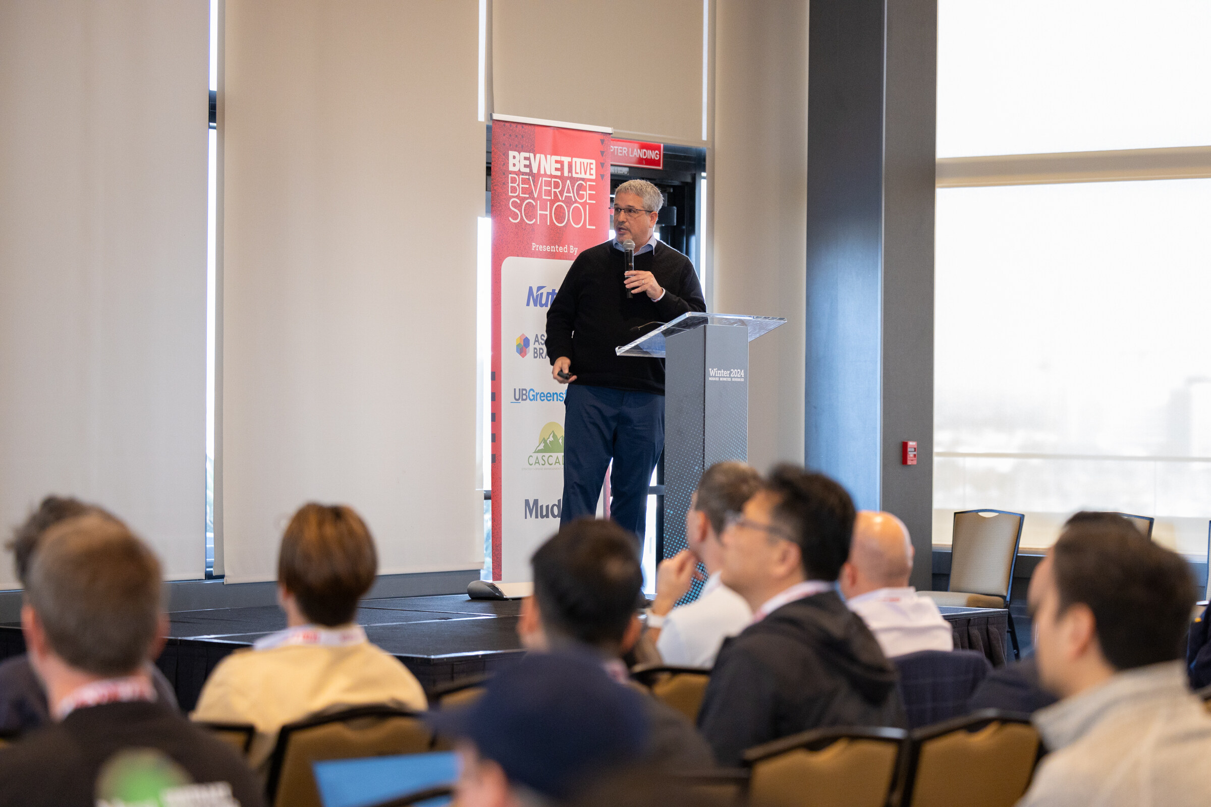 Rows of attendees face a man in dark clothes speaking at a lectern next to a Beverage School banner.