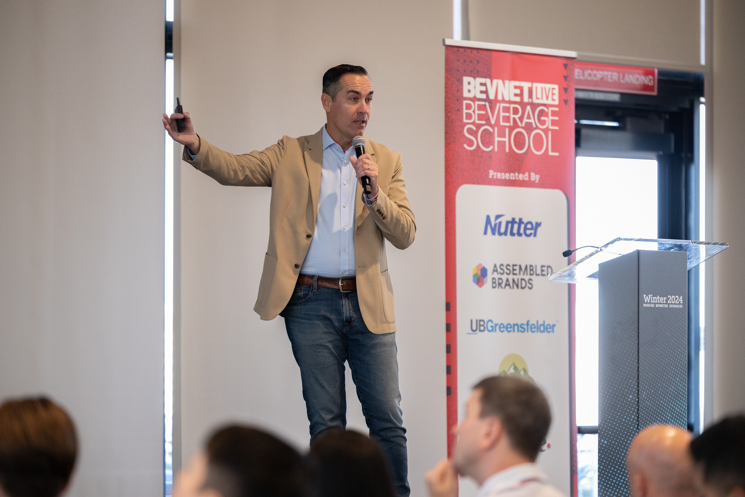 Man in a tan blazer and jeans points with one arm raised and holds a microphone beside a Beverage School banner.