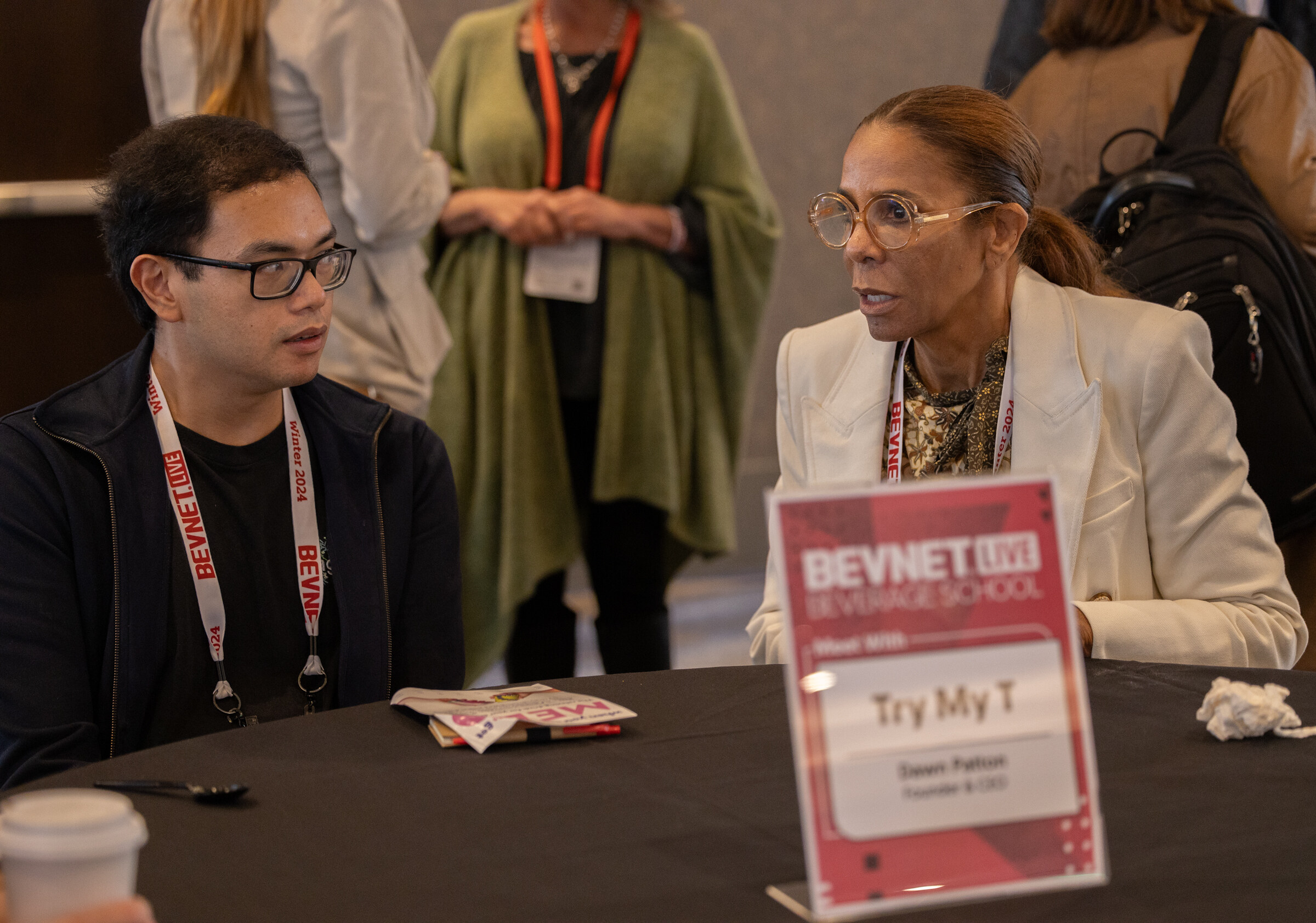 Two people with BevNET lanyards talk across a black table that has a Beverage School place card.