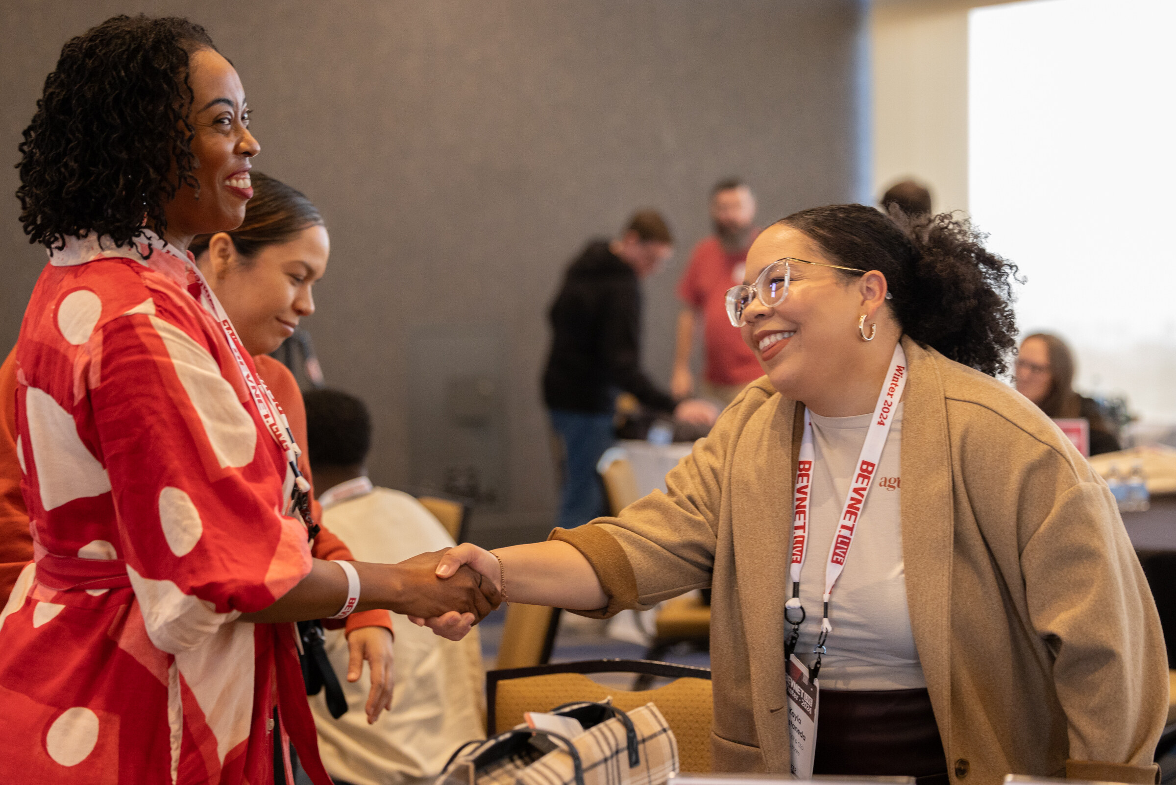 A woman in a red patterned dress shakes hands with a smiling woman in a camel coat.