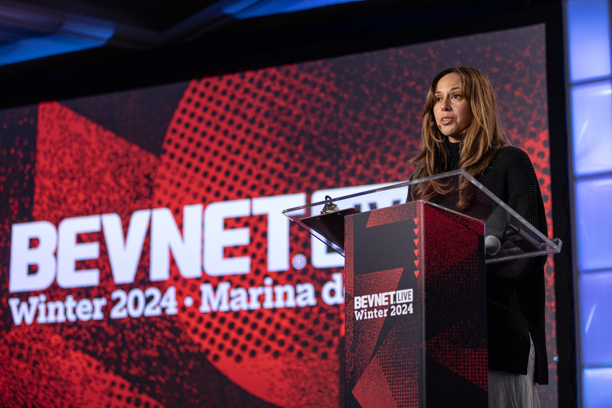 Woman with long hair in black speaks at a clear podium beside a BevNET Live Winter 2024 Marina del Rey screen.
