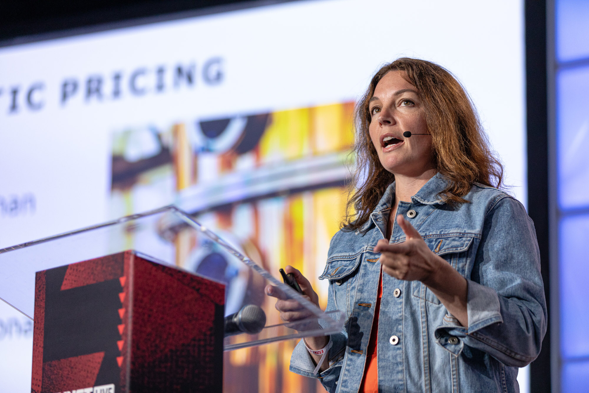 Woman in a denim jacket speaks at a podium with a slide that reads pricing behind her.