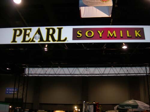 Wide overhead banner reading Pearl Soymilk in black and yellow lettering, above a truss structure in a dim exhibit hall.