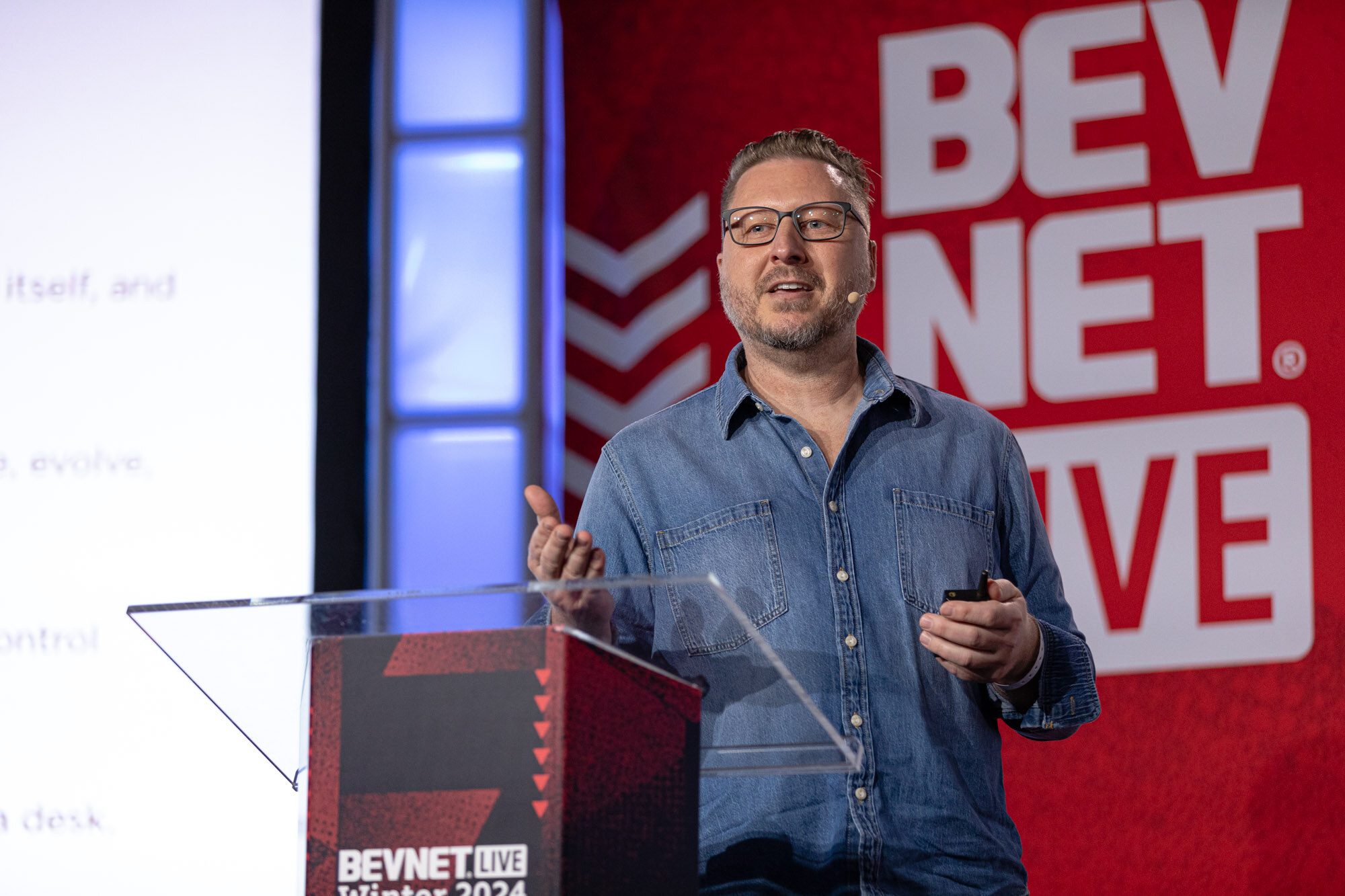 Man with glasses in a denim shirt stands at a podium and gestures in front of a BevNET Live backdrop.