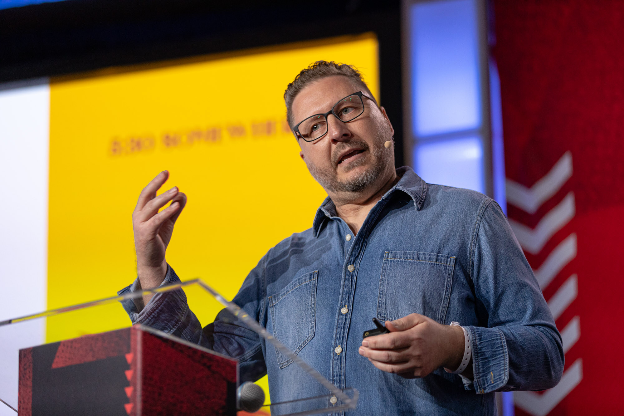Man in glasses and a denim shirt gestures with both hands at the podium.