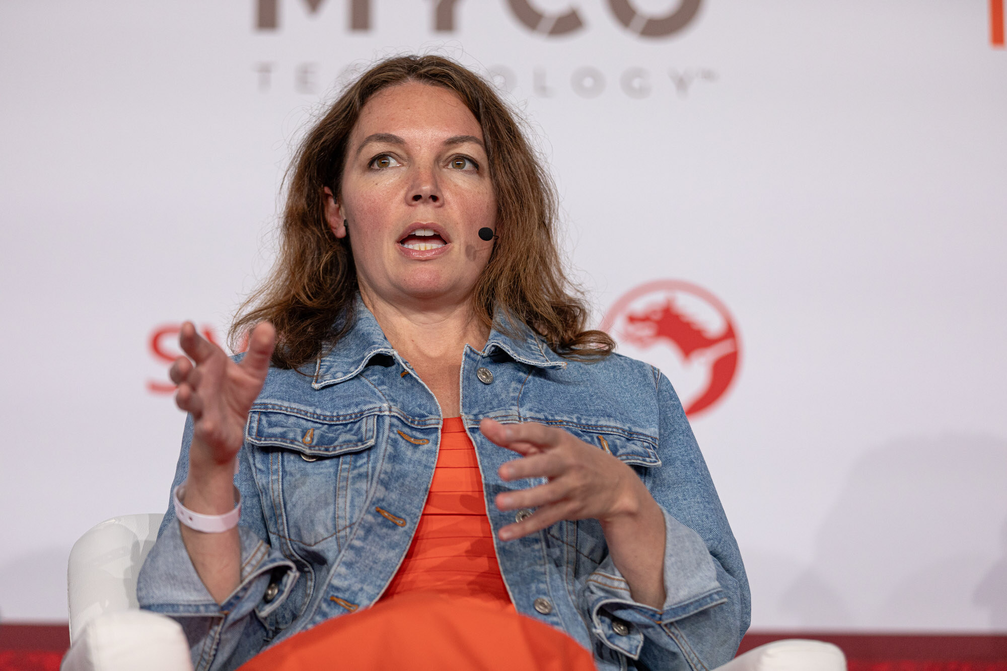 Woman in a denim jacket and orange top gestures while seated in front of a sponsor wall.