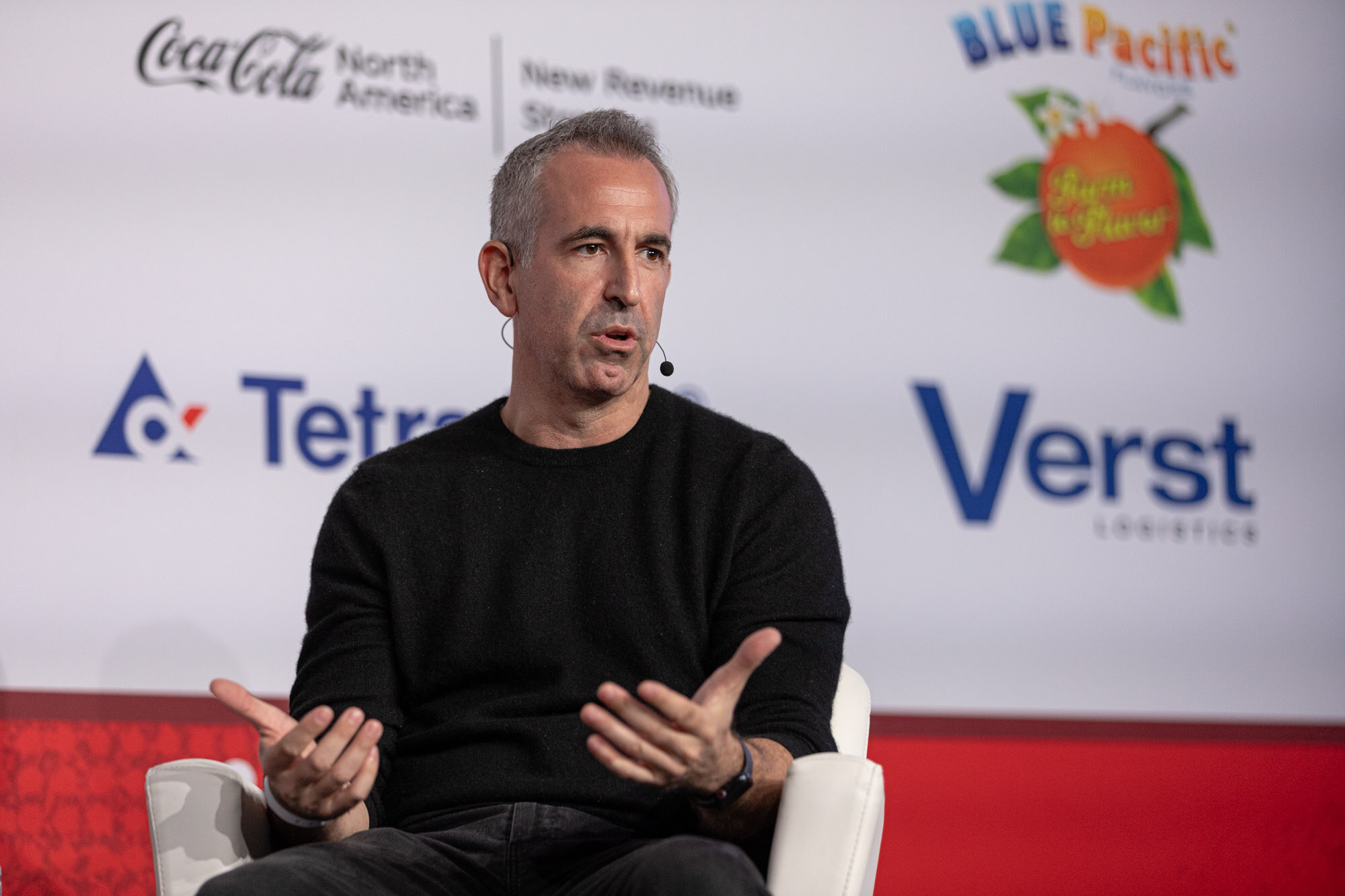 Man in a black sweater gestures with both hands while seated in front of a Coca-Cola, Tetra Pak and Verst sponsor wall.