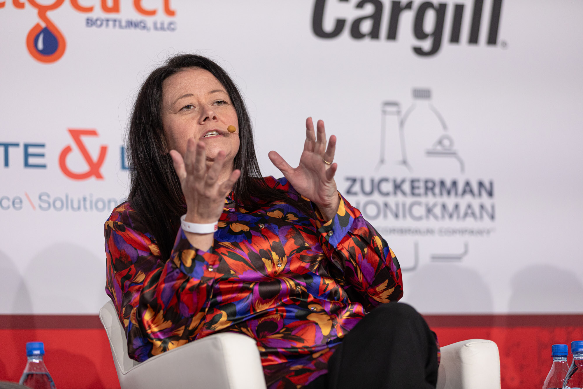 Woman in a bright patterned shirt raises both hands while seated in front of a sponsor wall.