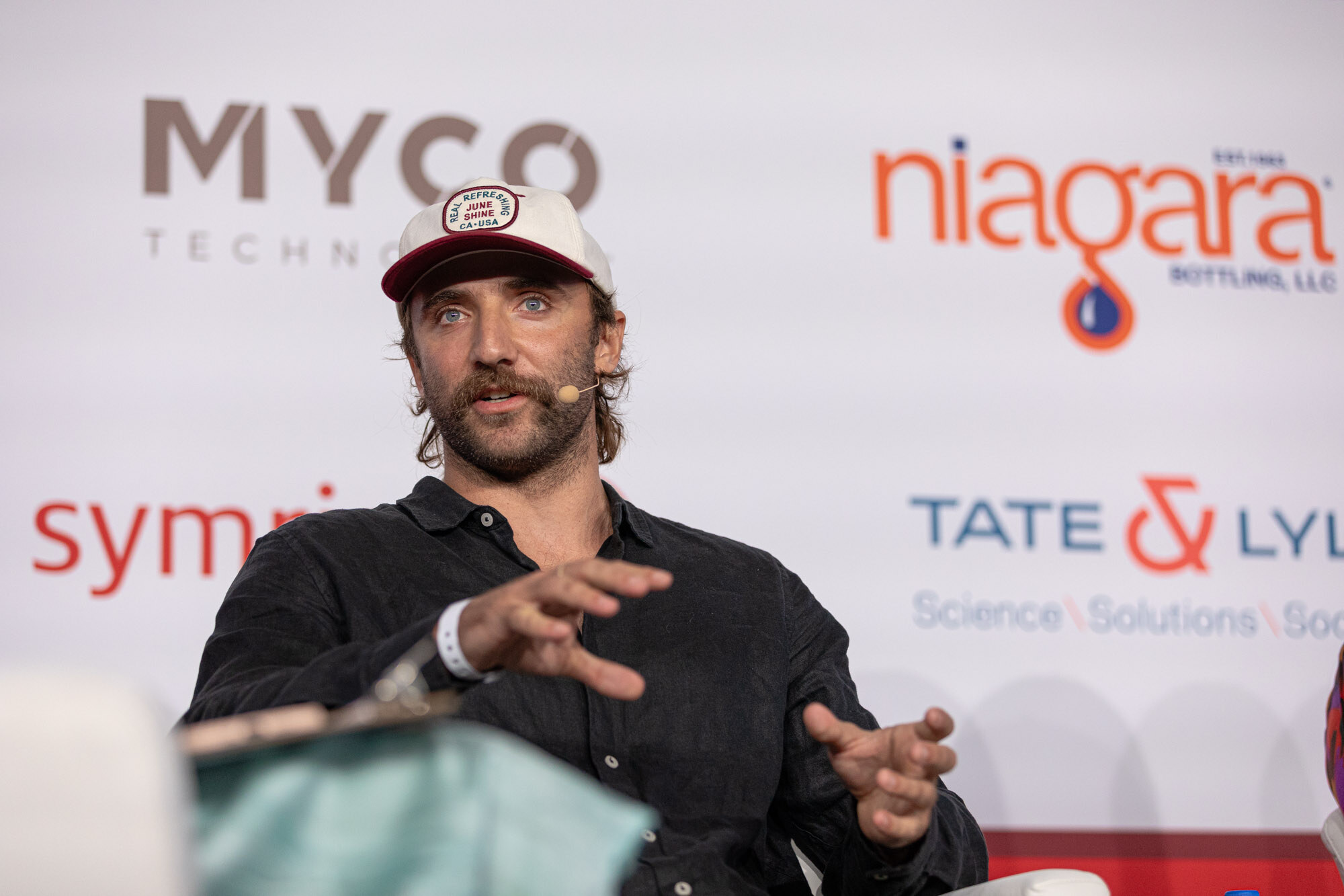 Man in a baseball cap and black shirt gestures while seated in front of a sponsor wall.