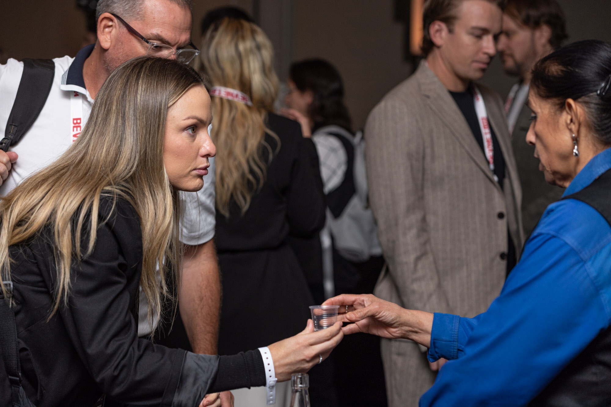 Several attendees stand in a crowd, one woman holds a small cup out to another.