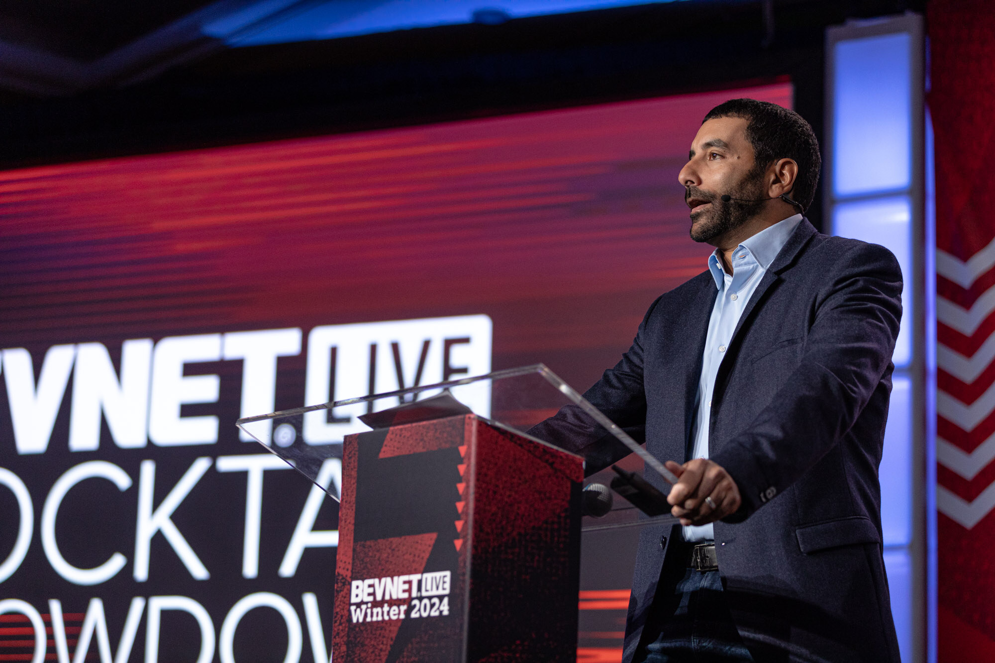 Man in a navy blazer speaks at a lectern in front of a screen showing part of the words Cocktail Showdown.