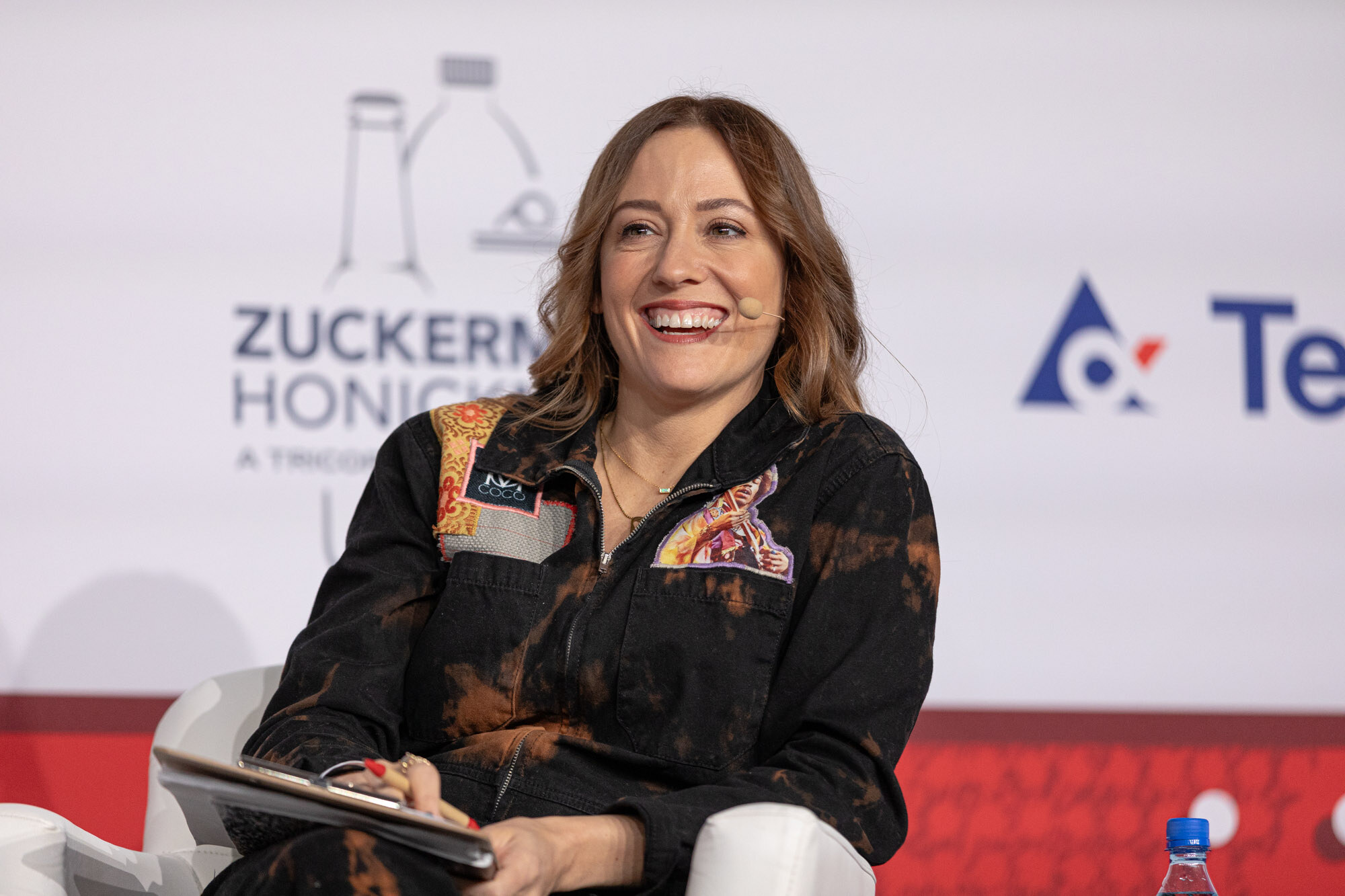 Smiling woman in a black patterned jacket sits in a white armchair in front of a sponsor wall.