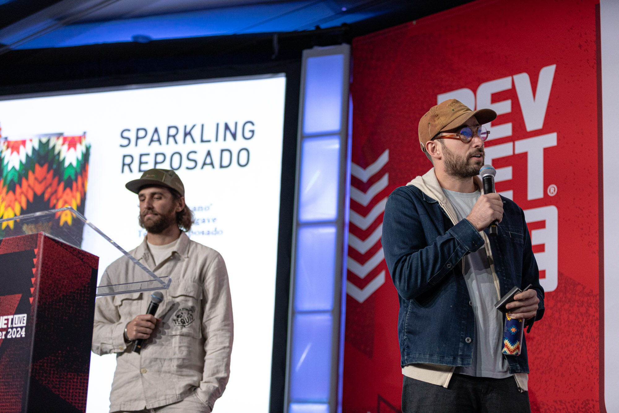 A man in a cap and cream jacket stands at a podium beside a man in a denim jacket in front of a Sparkling Reposado slide.