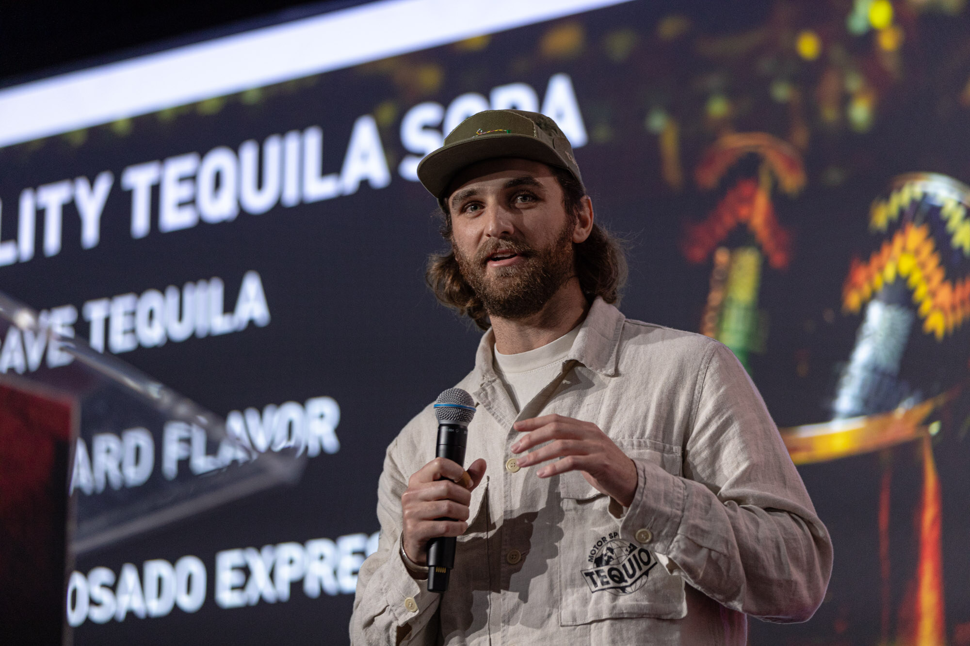 Bearded man in a green cap and cream jacket speaks into a microphone in front of a screen with tequila text.