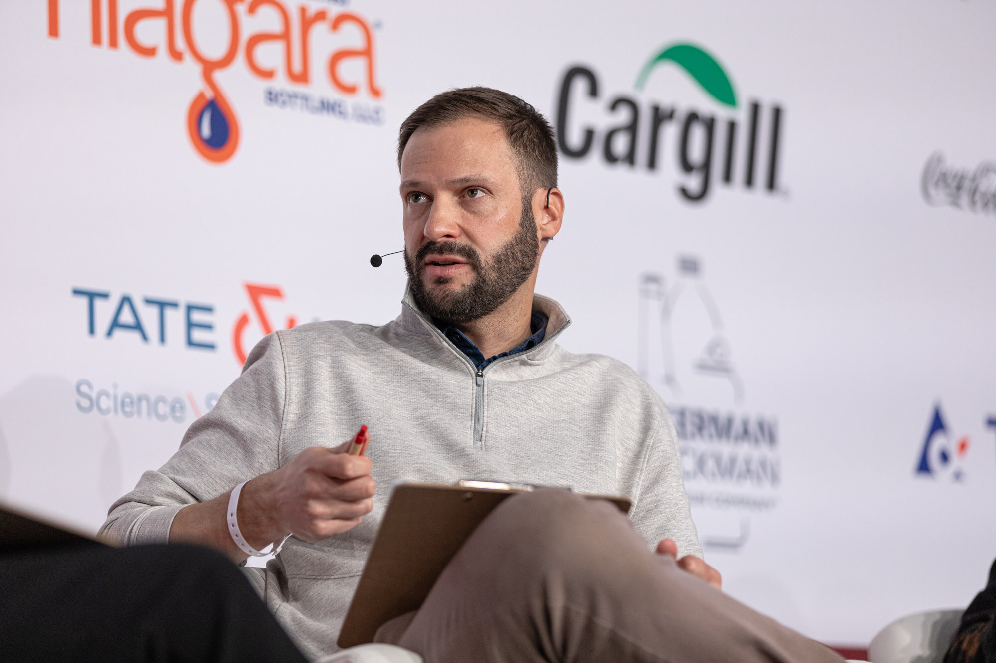 Bearded man in a gray quarter-zip holds a clipboard and a pen while seated on stage.