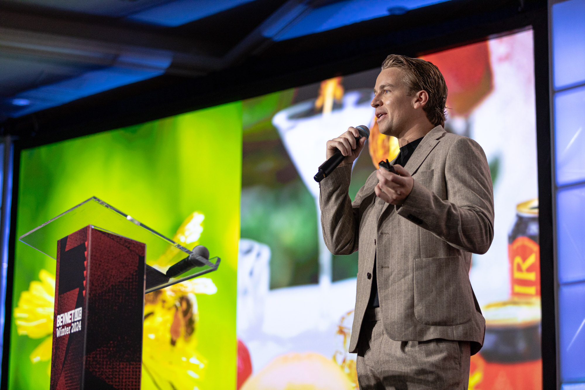 Man in a light tan suit holds a microphone in front of a large screen with flowers and a cocktail glass.