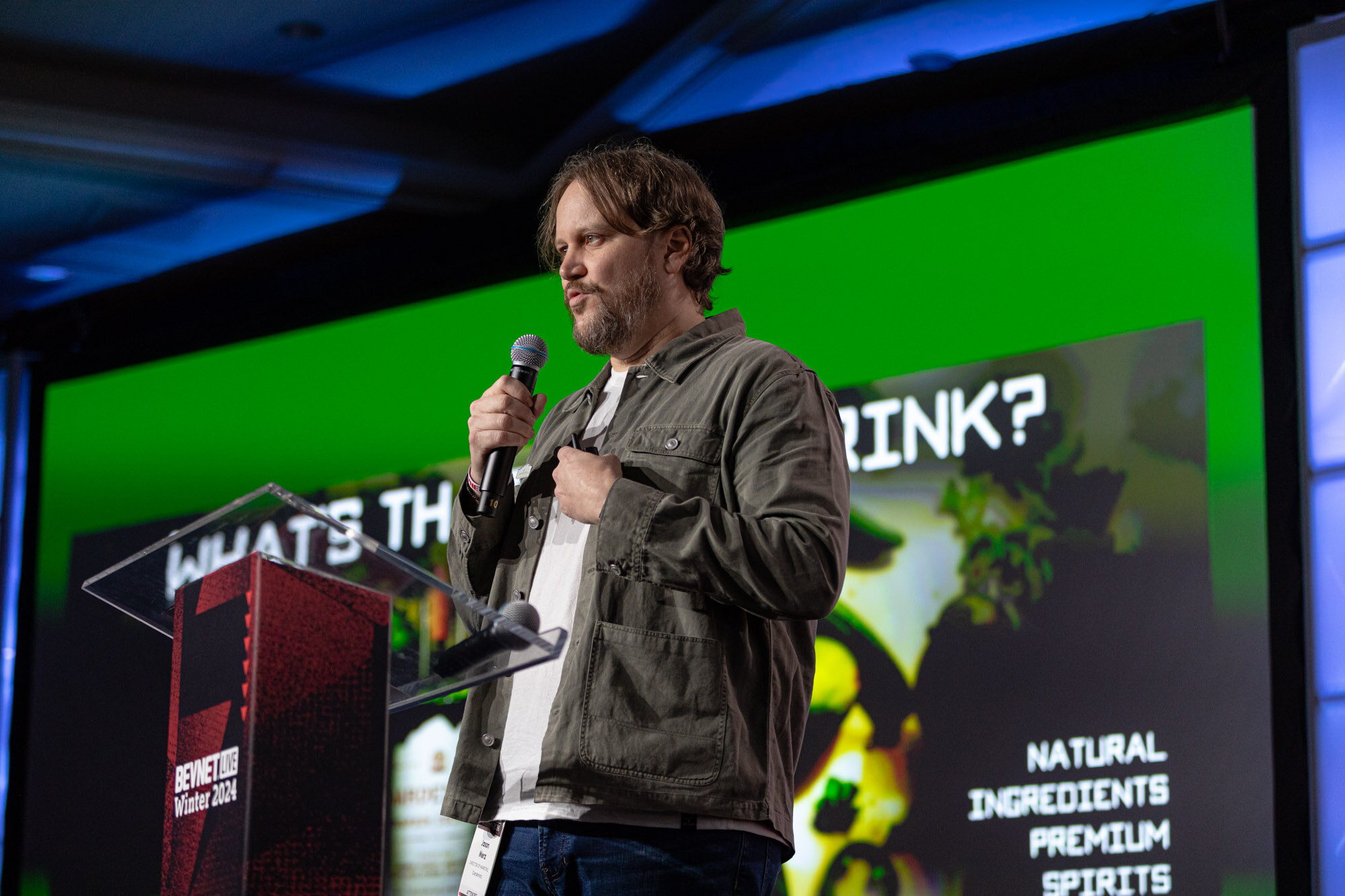 Bearded man in an olive jacket speaks in front of a green slide that reads natural ingredients premium spirits.