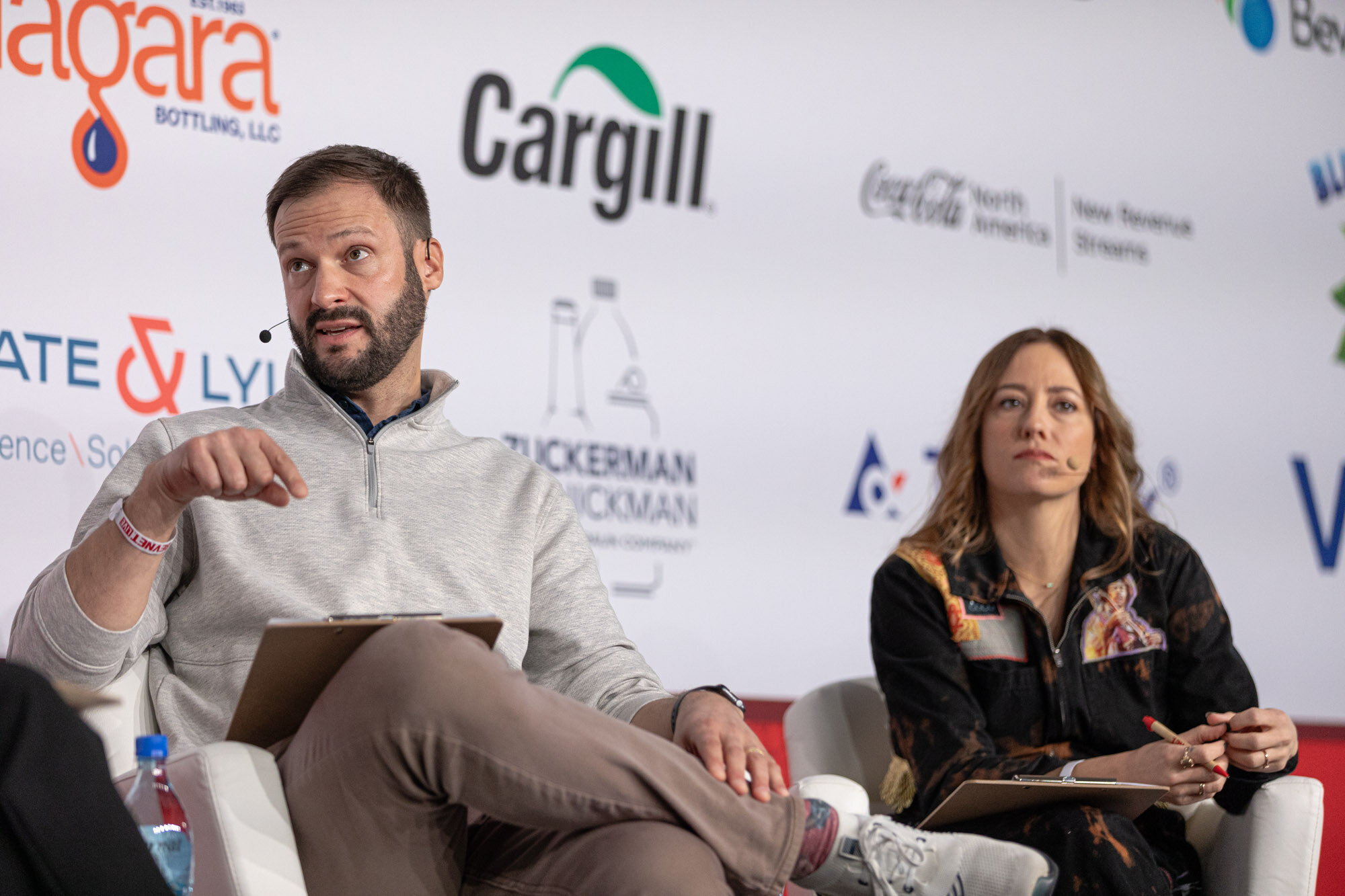 A bearded man with a clipboard and a woman in a patterned jacket sit side by side on stage.